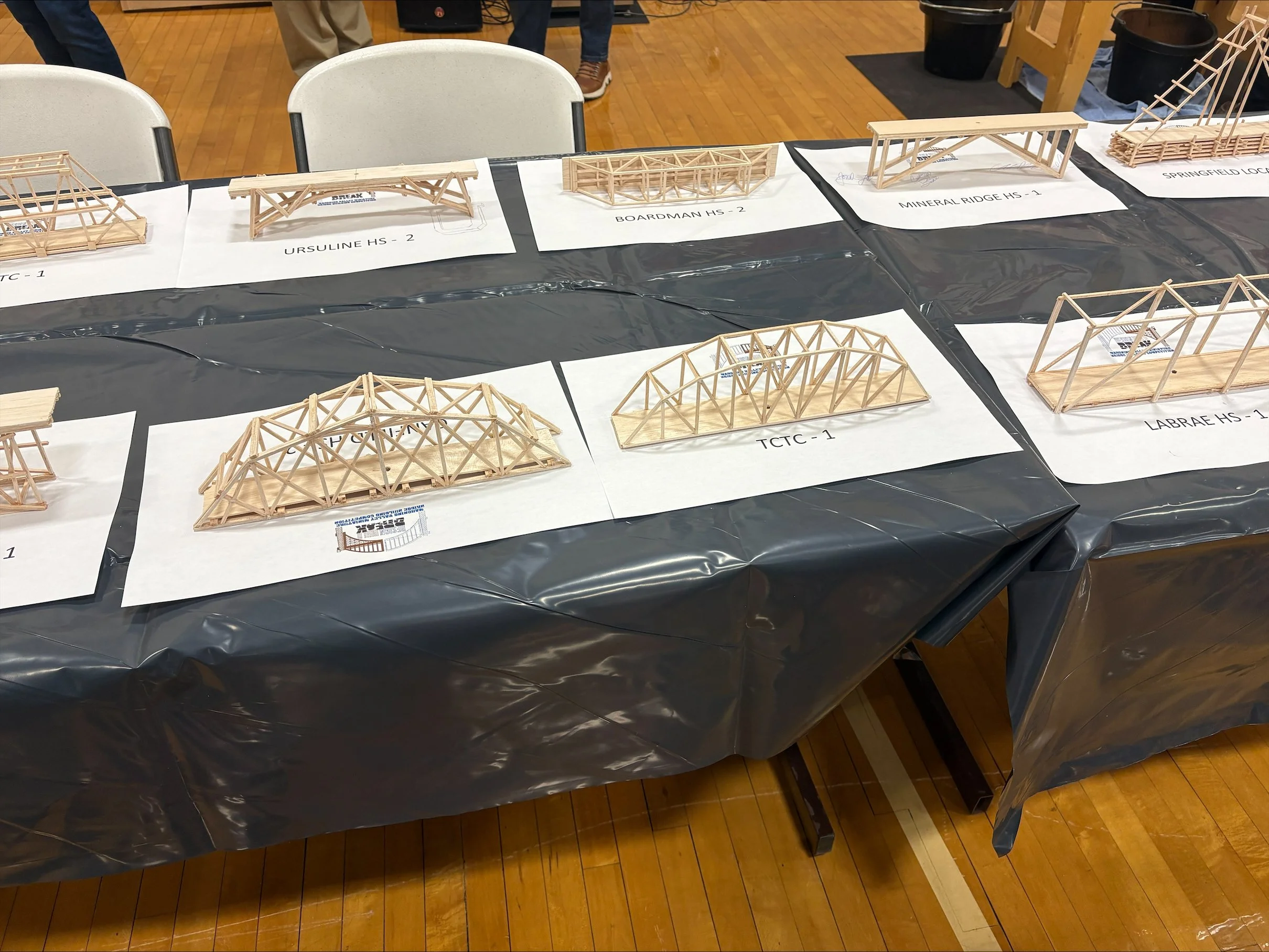 Display of miniature wooden bridges on a table at an event, each labeled with different school names.