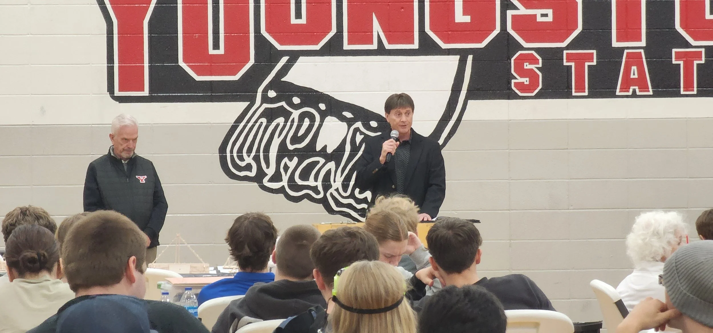 A man is speaking into a microphone at a crowded indoor event, with a large Yale Bulldogs logo painted on the wall behind him. Another man is standing to his side. Audience members are seated at tables.