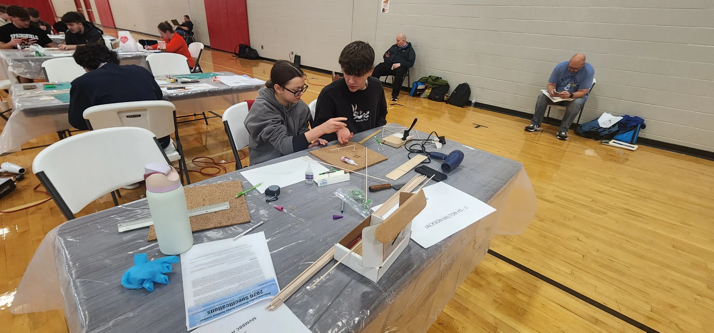 Two people are working on a project at a table with various tools and materials, while others in the background are sitting and standing in a gymnasium.