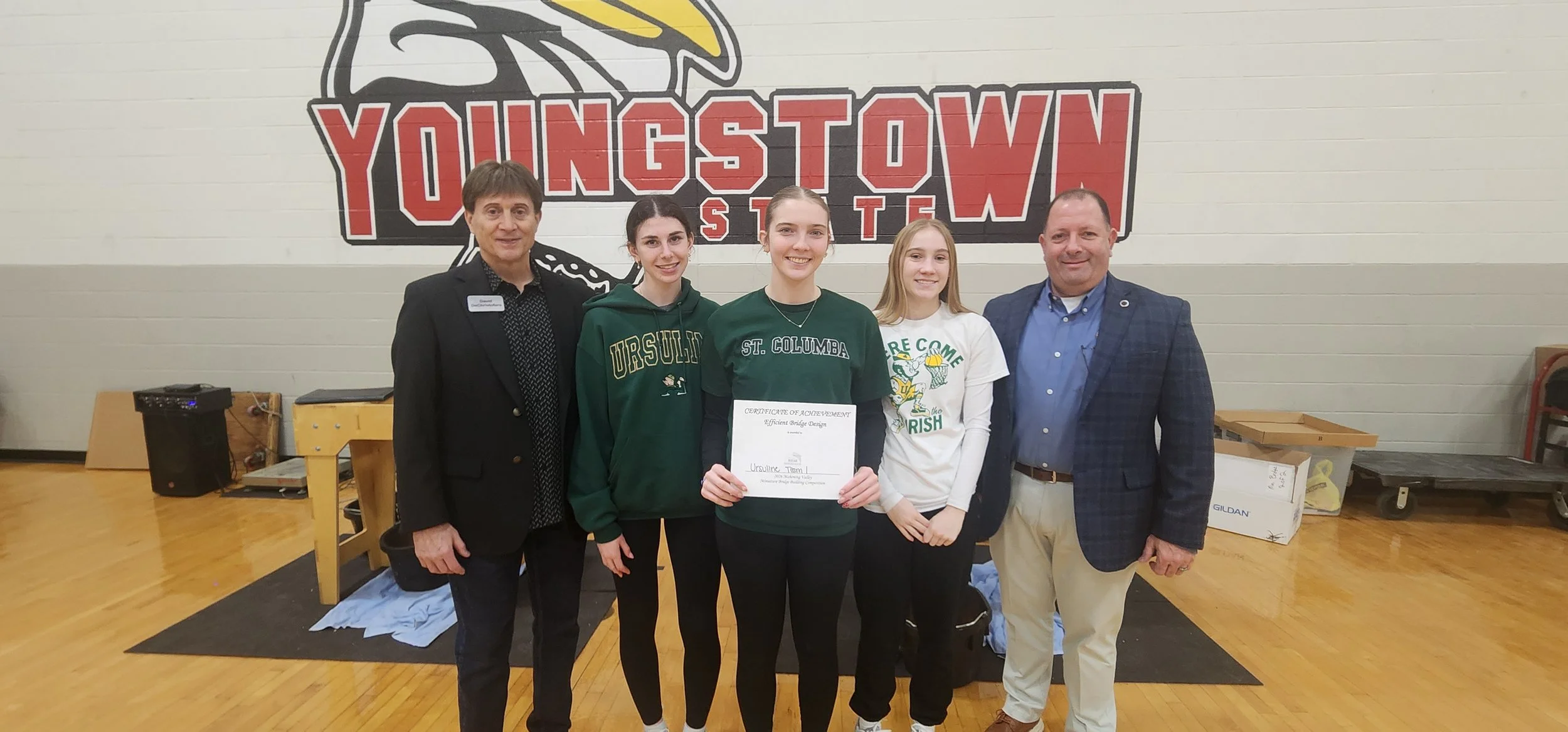 Five individuals stand in front of a large 'Youngstown State' logo on a gymnasium wall, with three females in the center holding a certificate of achievement. The individuals appear to be at an award ceremony or school event.