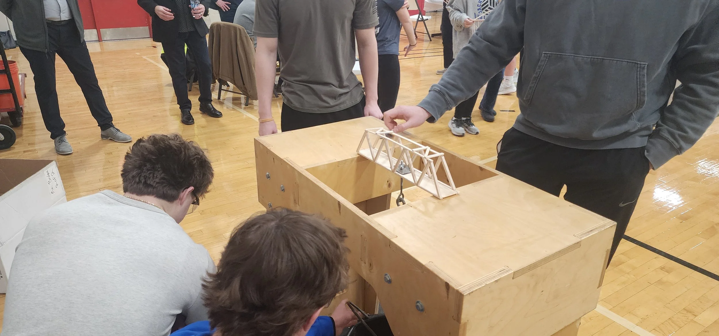 Multiple people gathered around a wooden table in a gymnasium, observing a small bridge model made of wooden sticks. One person is pointing at the model, while others are watching or kneeling nearby.