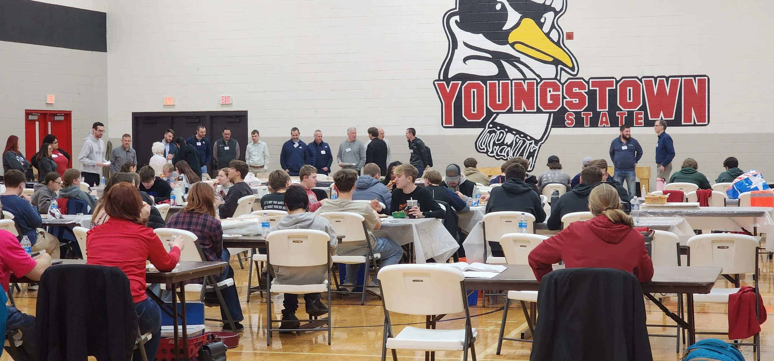 A group of people gathered in a large indoor gymnasium, sitting at tables and listening to a panel of speakers standing in front of a wall with a large graphic of a duck and the words "Youngstown State".