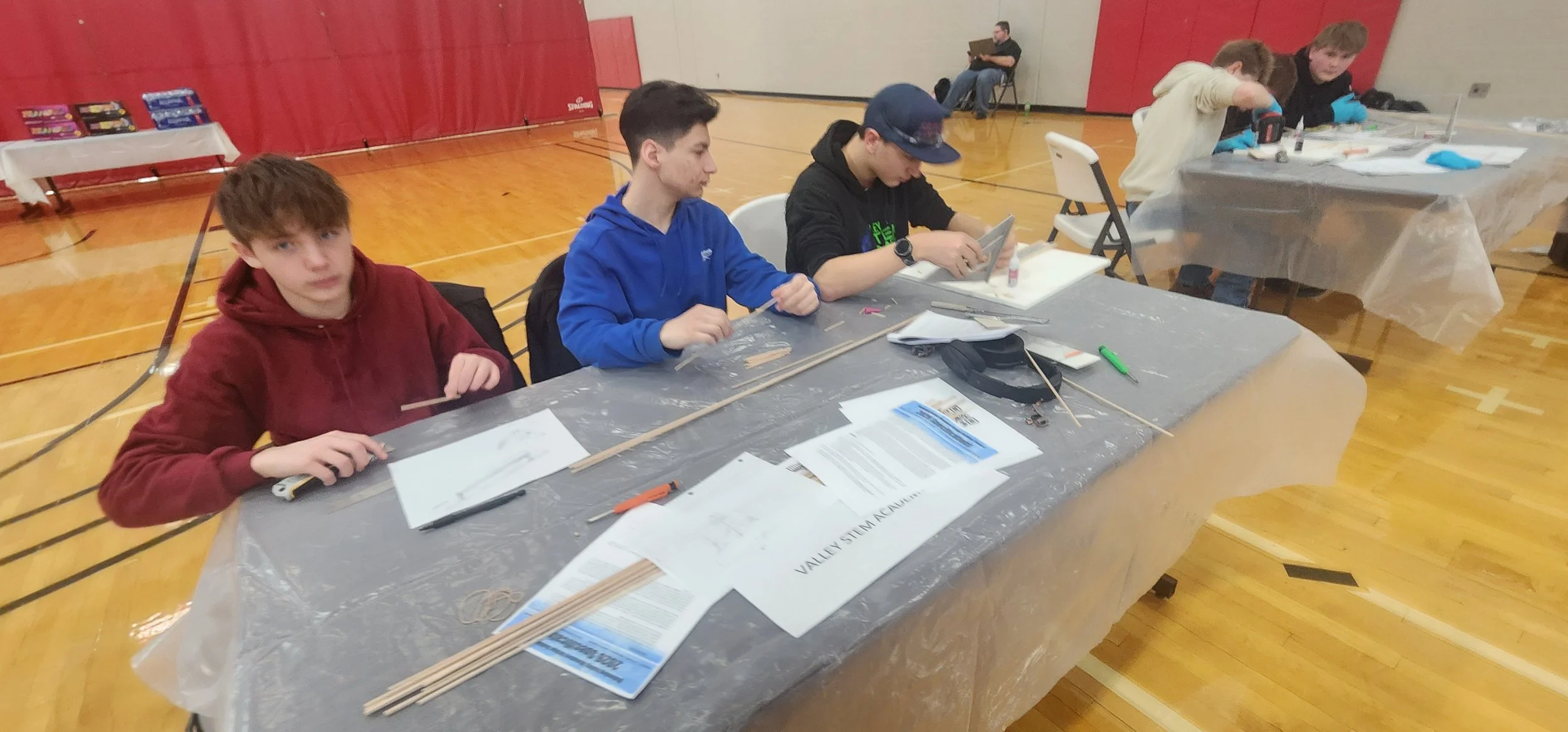 A group of students working on a project at a table in a gymnasium, with some building supplies and documents scattered on the table.