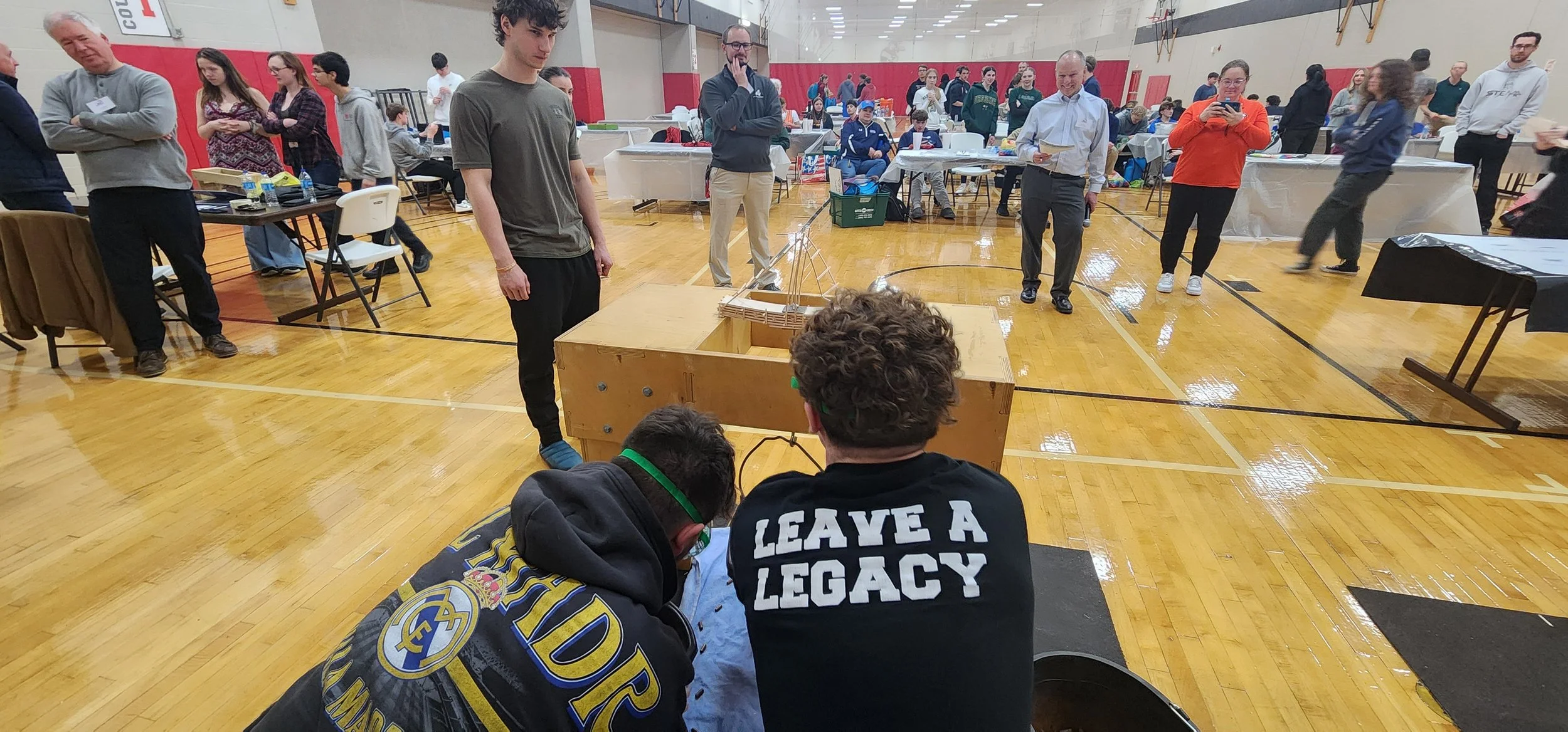 People gathered in a gymnasium watching a demonstration involving a small robotic vehicle on a table. Some are taking photos or notes, and others are observing.