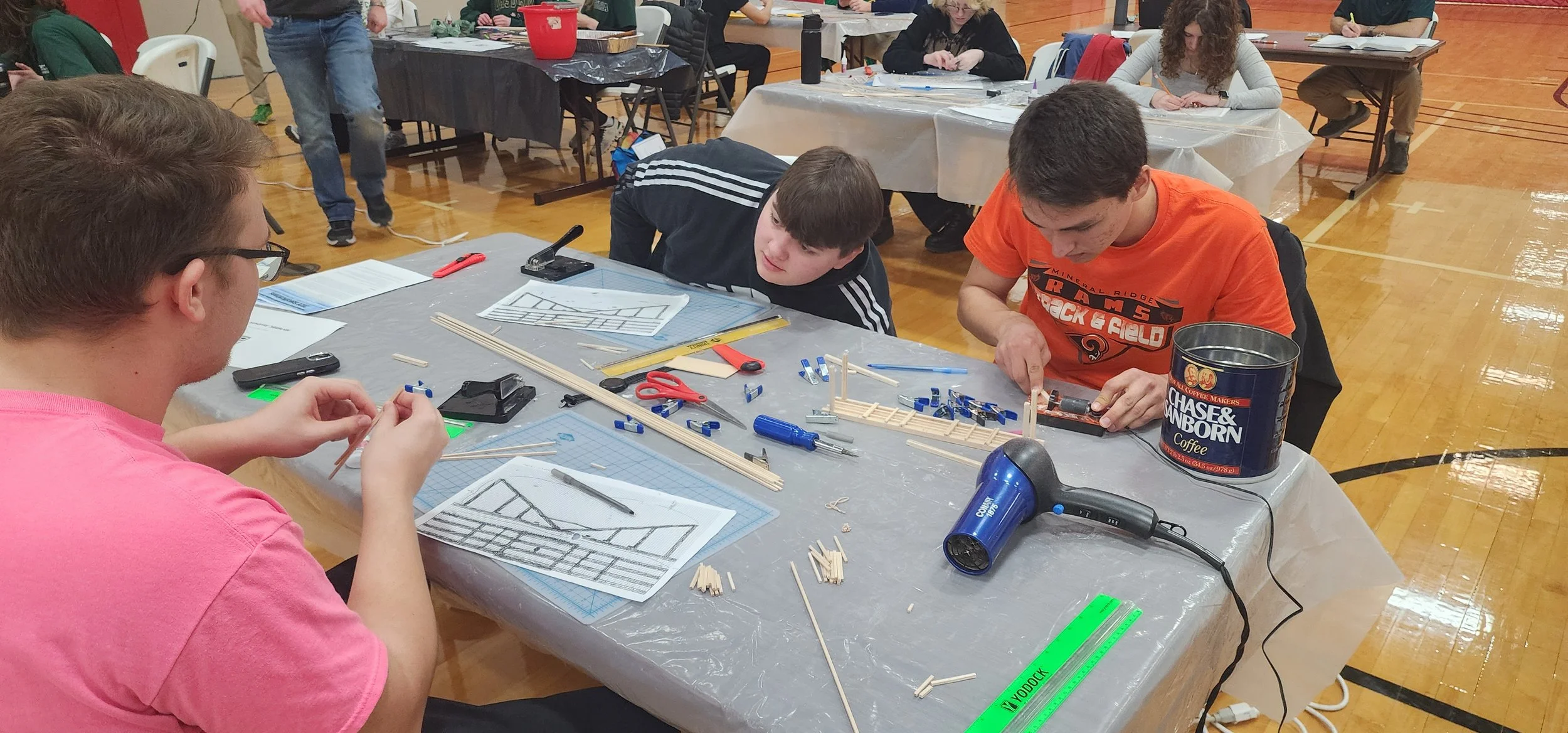 Three young boys working on a model-building project at a table in a gymnasium. The table is covered with tools, rulers, and small wooden pieces. Other people are working on similar projects in the background.