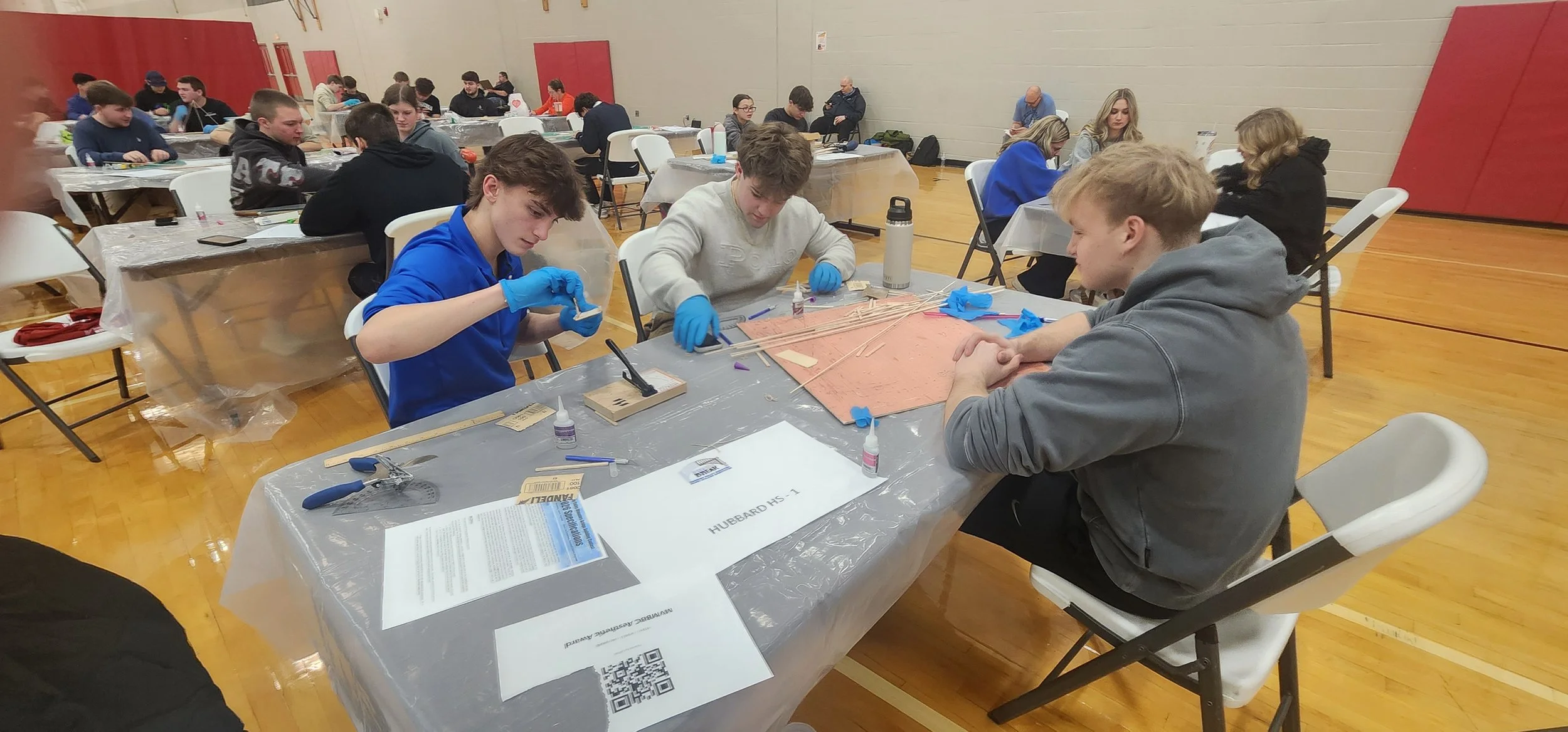 Several students working on a project at a table in a school gymnasium with banners on the walls.