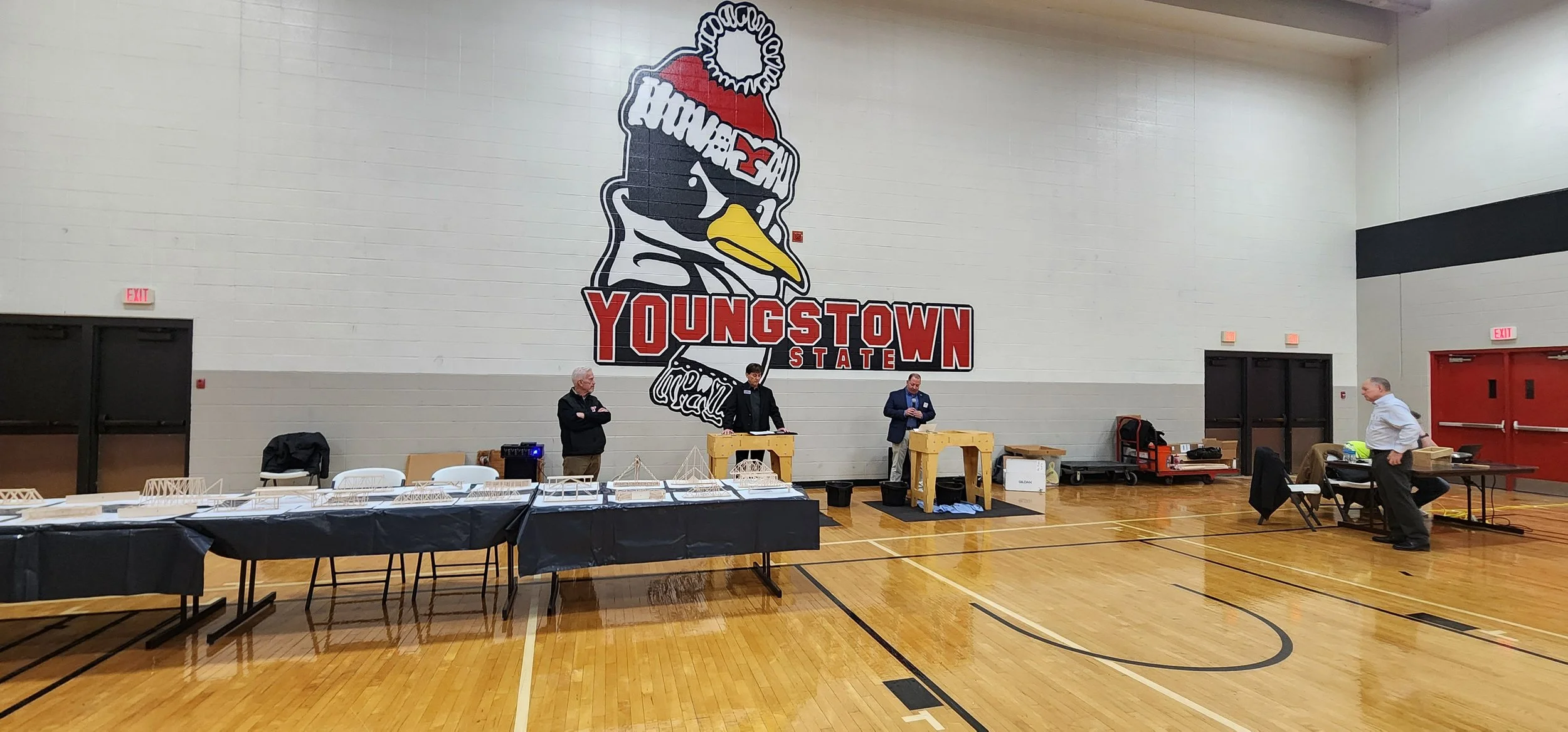 Indoor gymnasium with a large wall mural of a cartoon duck wearing a red beanie hat and sunglasses, with the words "YOUNGSTOWN STATE" below. There are three men standing near the wall, and tables with various items set up along the floor.