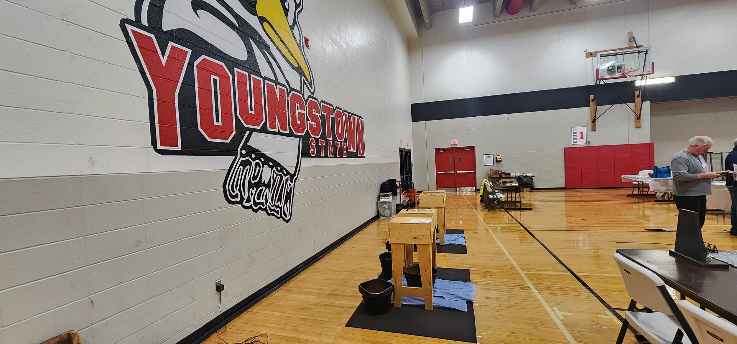 An indoor basketball gymnasium with a large Oklahoma State logo painted on the wall. There's a basketball hoop mounted on the wall, tables with supplies, and a man standing near the tables reading a paper or device.
