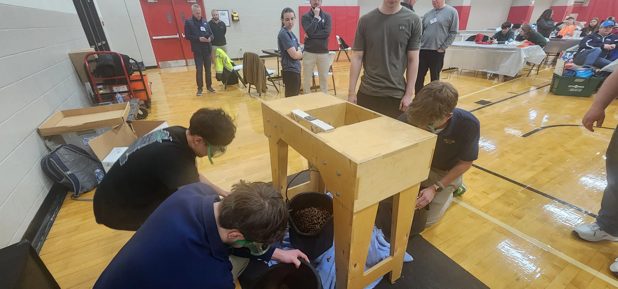 Group of students working together on a project in a school gymnasium, some kneeling around a wooden machine with a bucket of small objects, others observing or standing nearby.