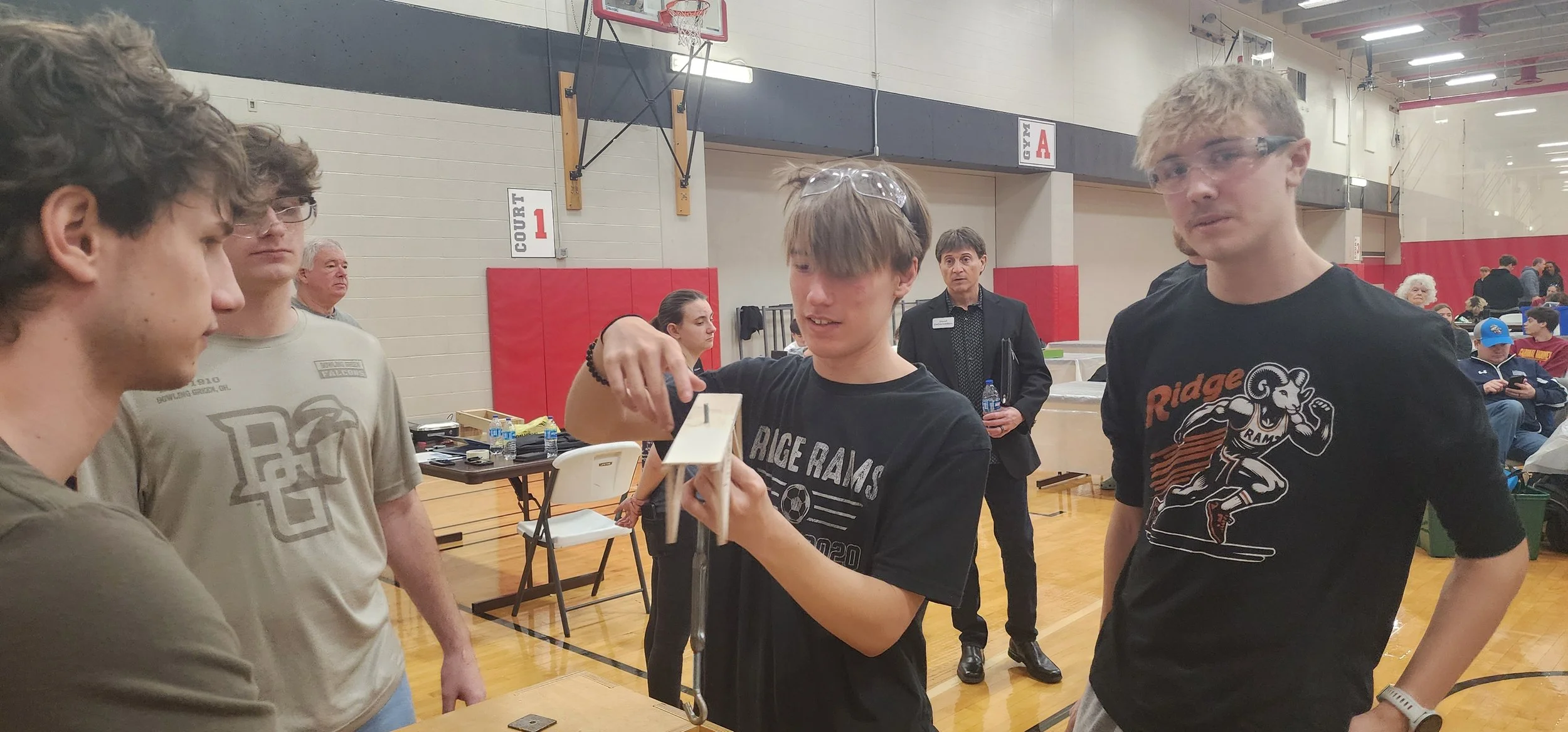 A group of four young males in a gymnasium, with some people seated in the background. One of the young men is holding and adjusting a small mechanical device, while the others watch him. They are wearing casual athletic clothing, and there are table