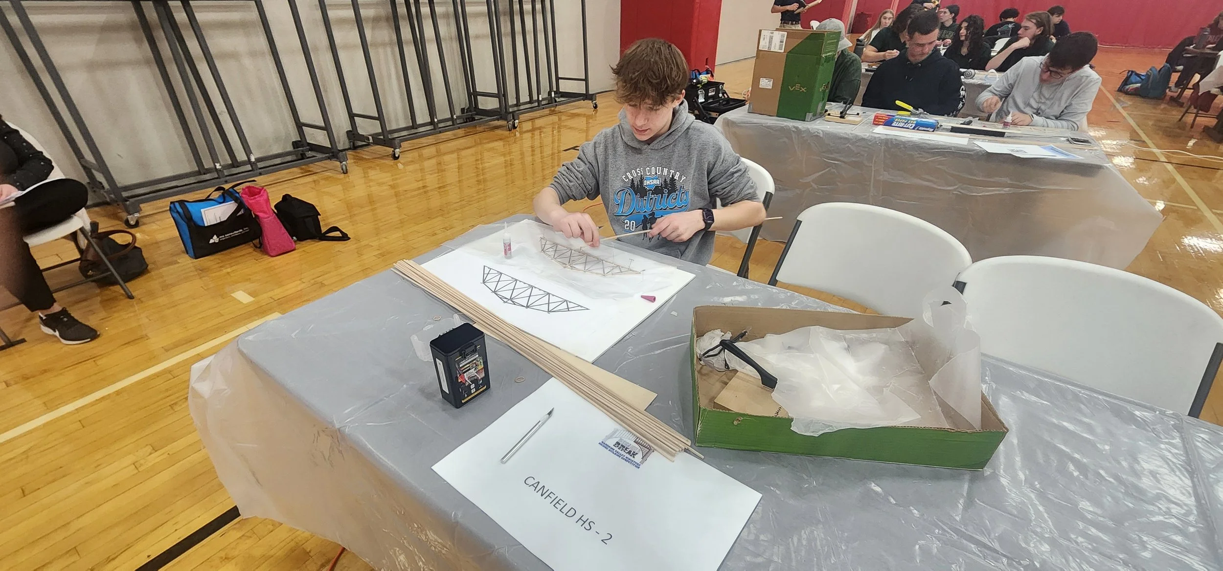 A young person working on a model bridge at a table in a gymnasium or hall during a robotics event, with other participants in the background.