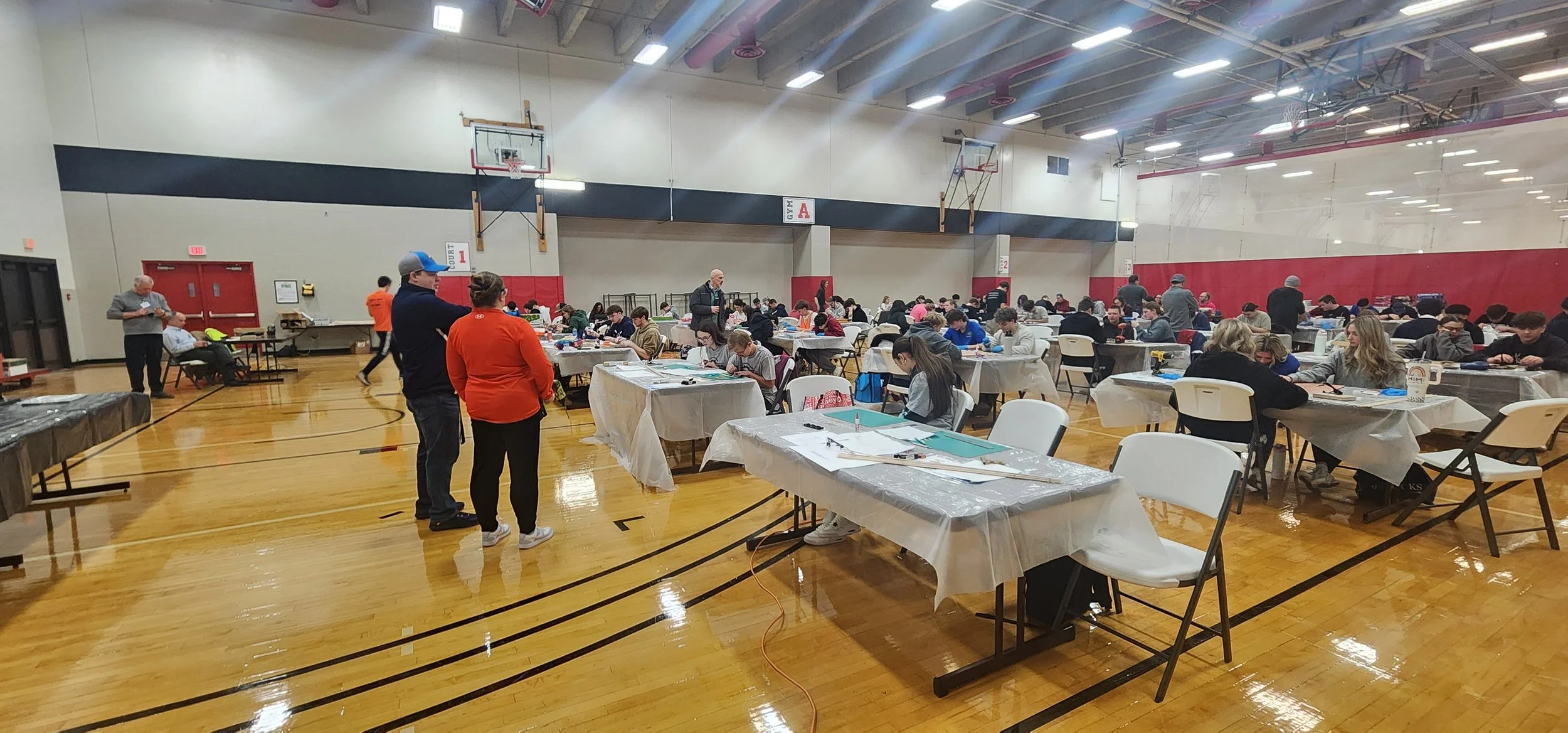 A large gymnasium filled with students seated at tables, taking a test or working on assignments, with some standing and talking. The gym has a wooden floor, basketball hoops, and a high ceiling with lighting.