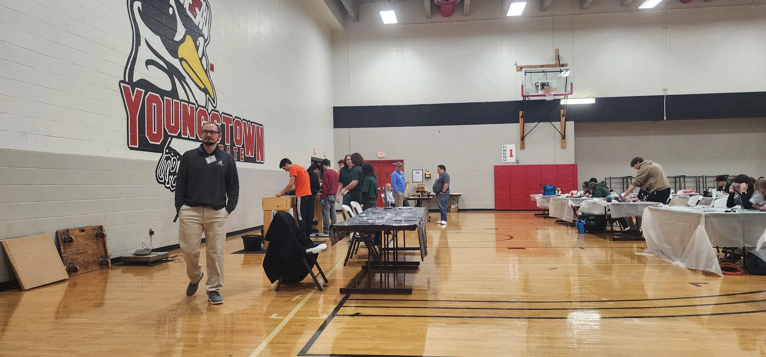 People gathered inside a gymnasium with tables set up, some working on crafts or projects, and a large logo on the wall for Youngstown State University.