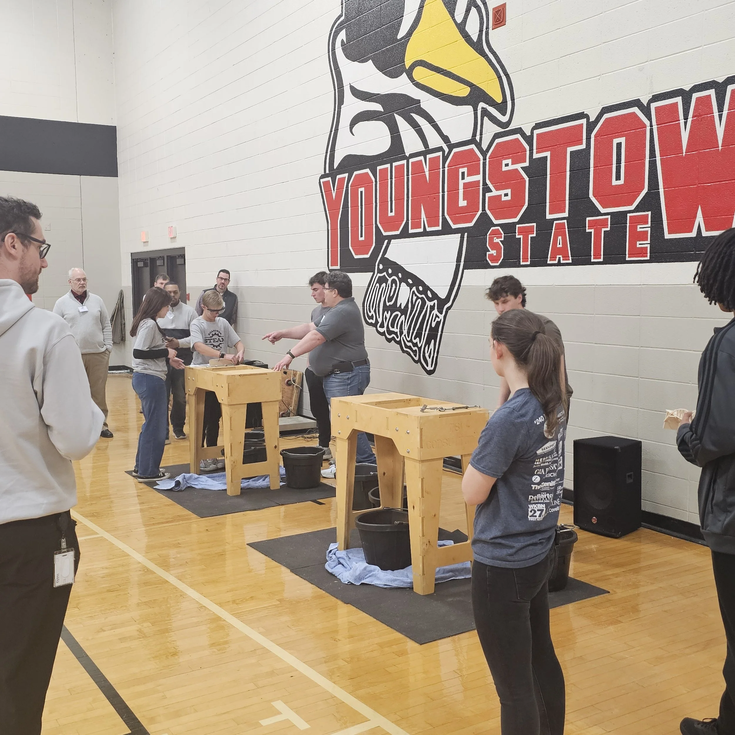 A group of people gathered in a gymnasium with a large mural on the wall featuring an eagle and the words "YOUNGSTOWN STATE" in red lettering. Some individuals are engaged in activities at wooden tables, and others are observing or talking. The room 