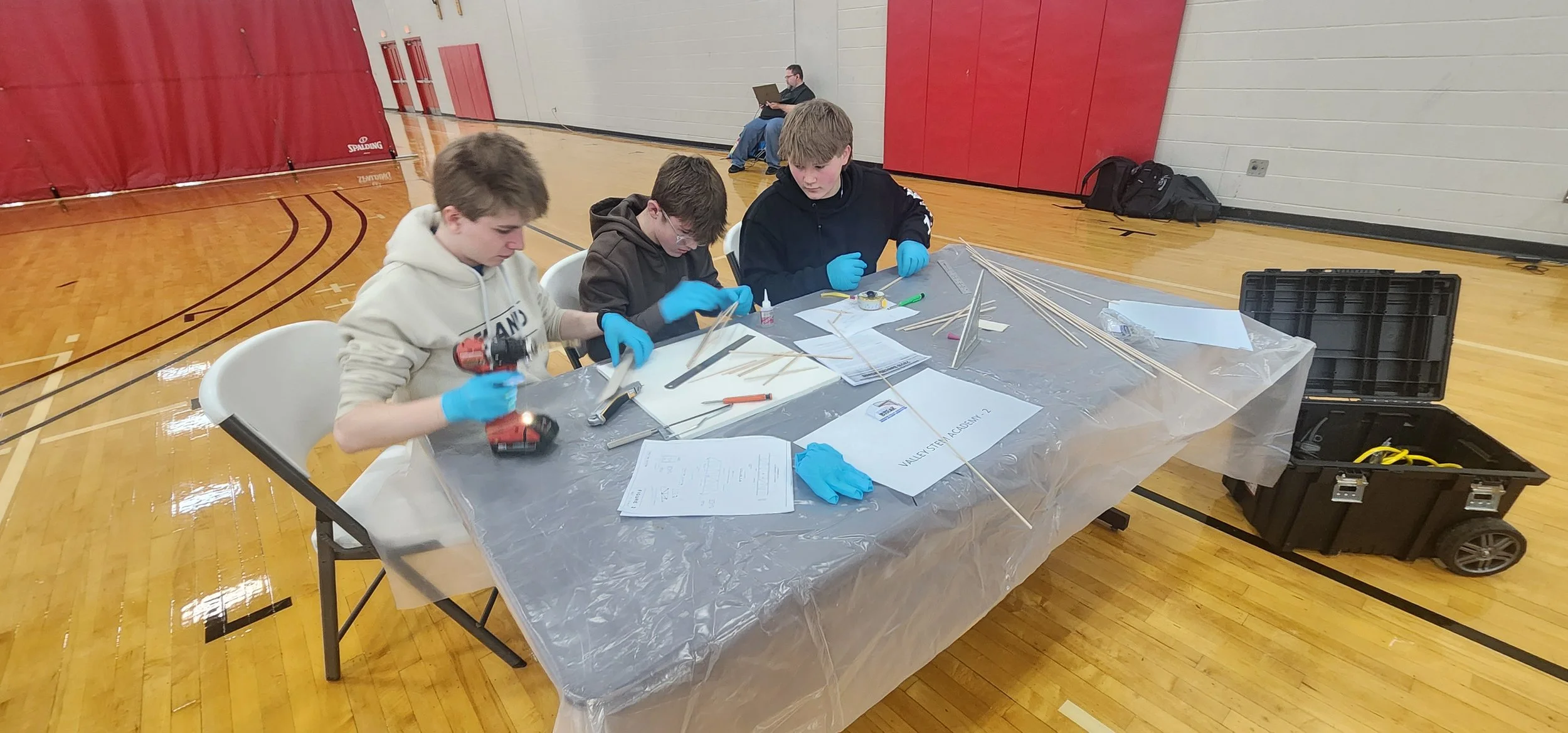 A group of three young boys sitting at a table in a gymnasium, working on a project with wooden sticks, tools, and papers. A supervising adult is in the background. The table is covered with a plastic sheet, and there is a black toolbox on the floor 