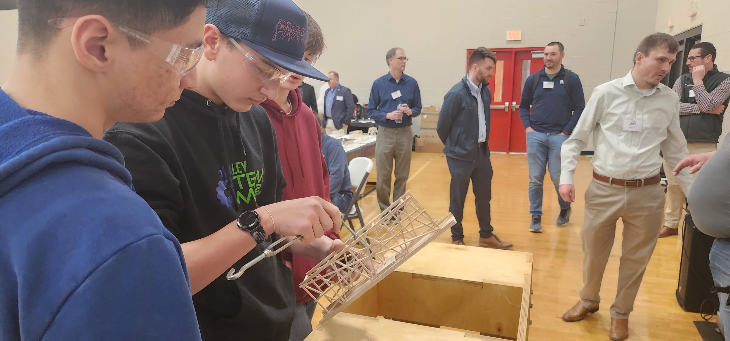 Young man wearing glasses and a smartwatch examining a small wooden bridge model in a busy indoor event.