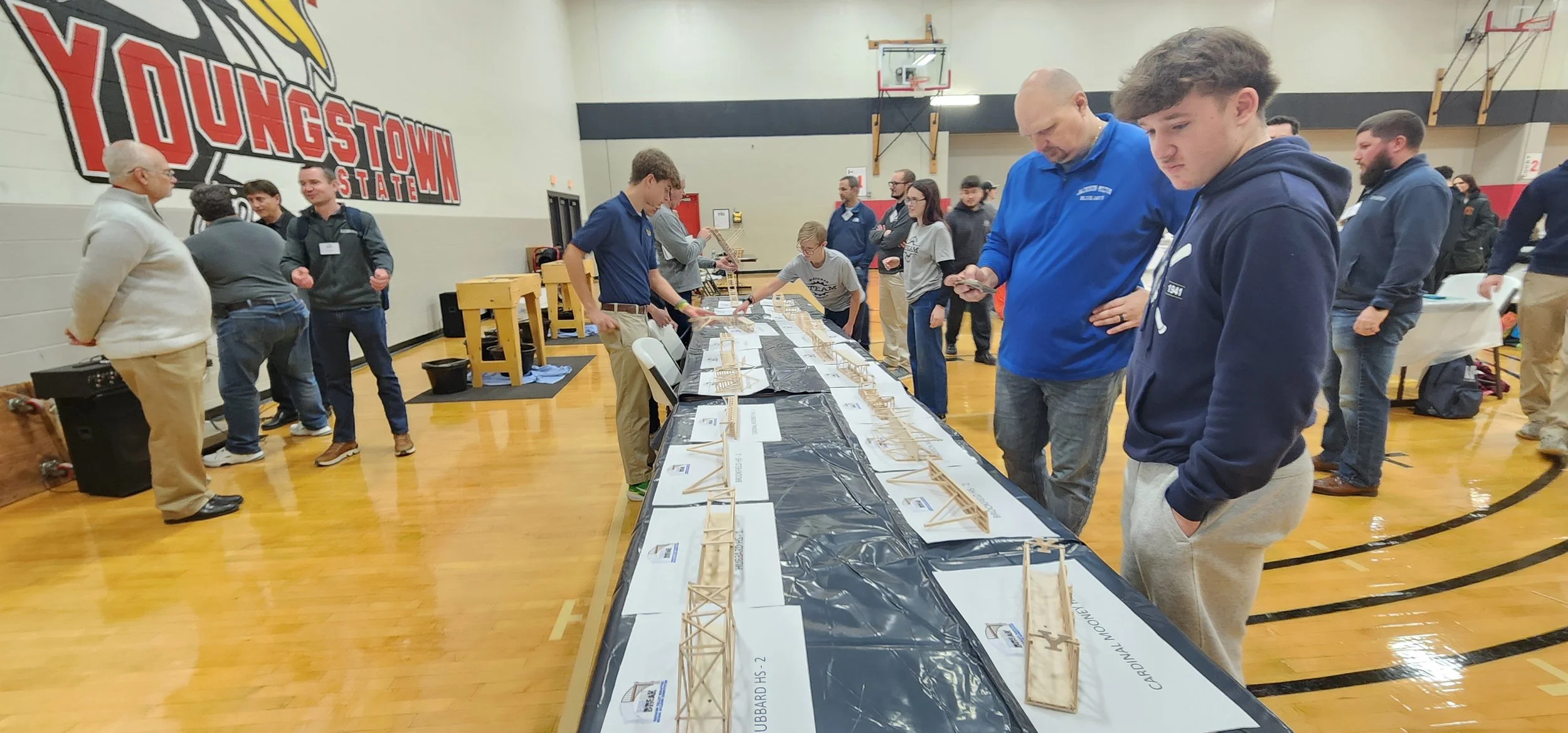 People standing around a table viewing model bridges in a gymnasium with a gymnasium wall and basketball hoop in the background.