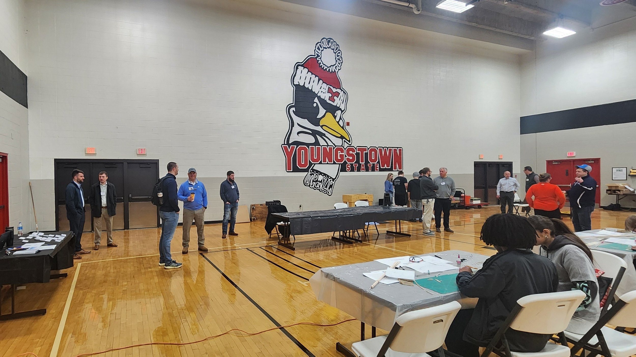 People gathering in a gymnasium with a large wall logo that reads 'YOUNGSTOWN STATE' and features a bird wearing a red and white hat, in a casual indoor event setting.