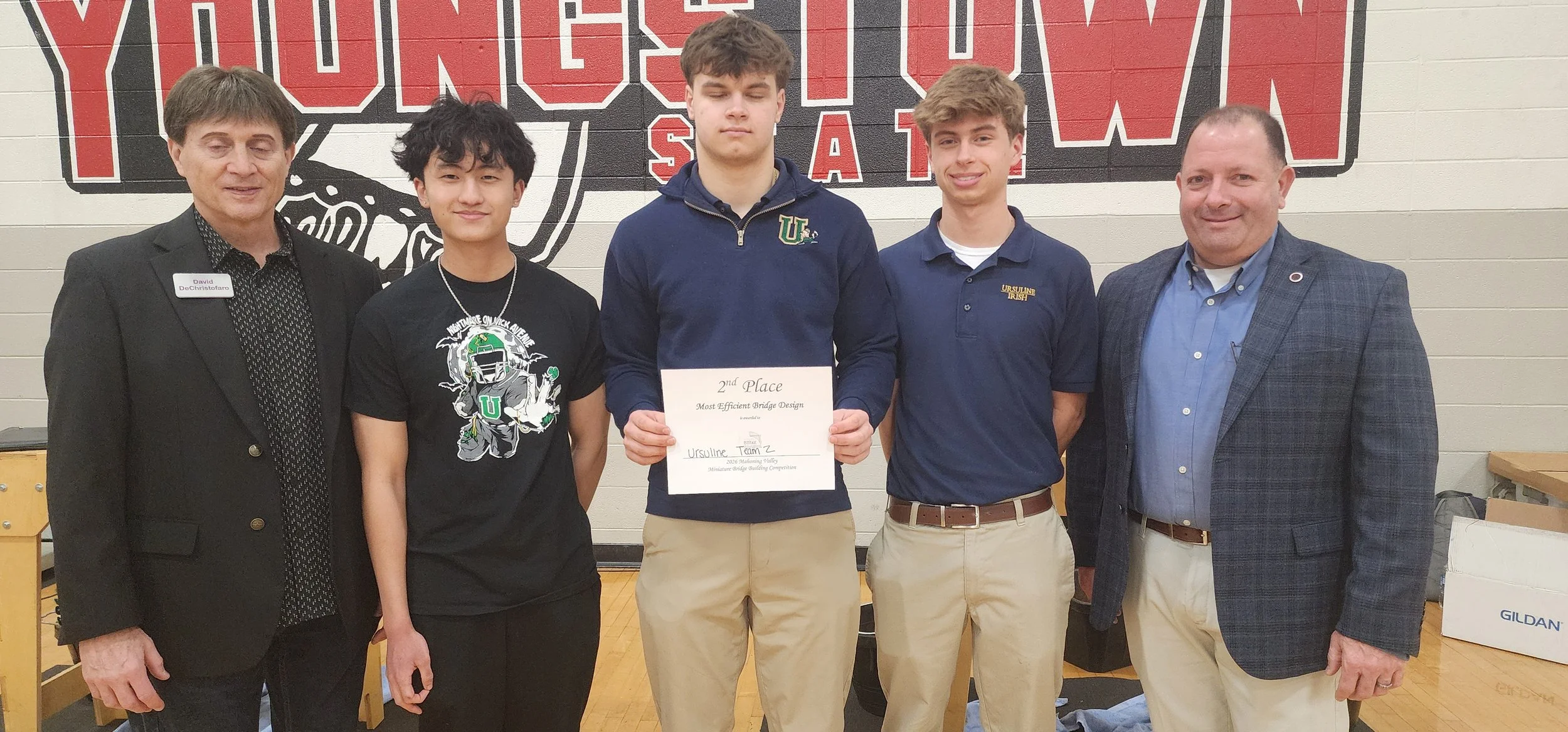 A group of five men and boys standing together in a gymnasium, with three of them holding a certificate for second place in a bridge building competition. The background features a gym wall with a large logo.