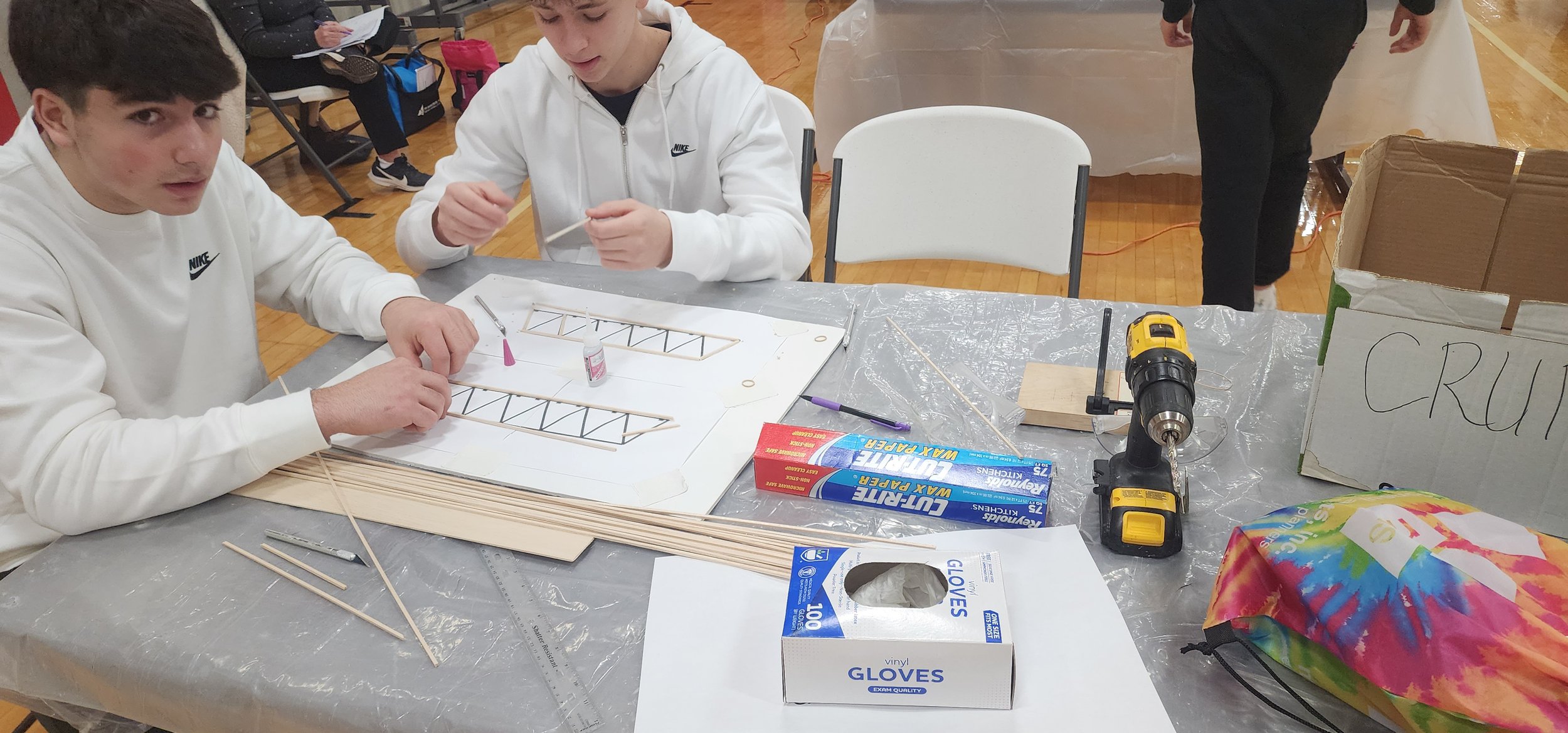 Two young men in white sweatshirts working on a model airplane at a table with tools, glue, and wooden sticks, in a gymnasium or large indoor space.