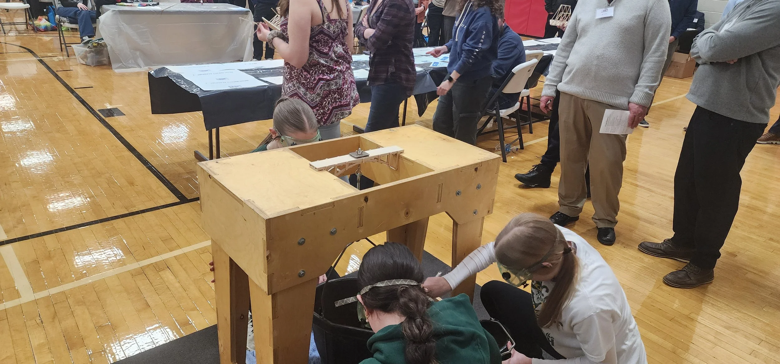 Children and adults gather around a small wooden robot in the center of a gymnasium during a robotics event.