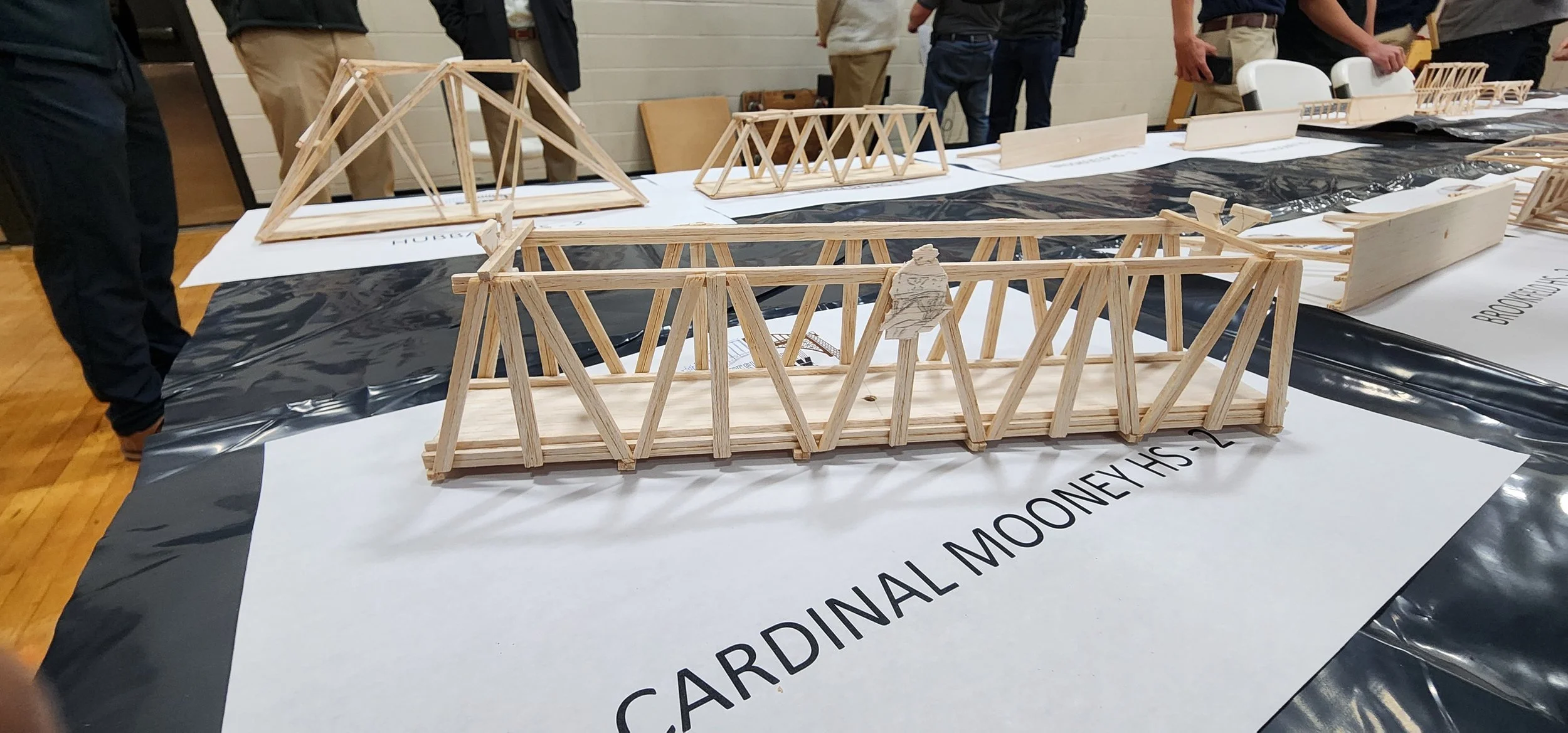 Wooden model bridges displayed on a table at an engineering or design event with people in the background.