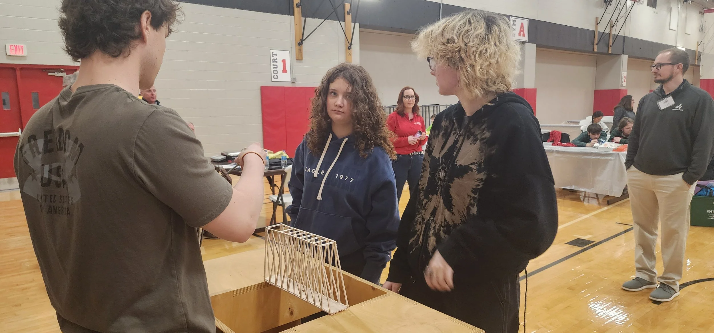 People in a gymnasium gathered around a table with a wooden bridge model, engaging in a discussion or activity.