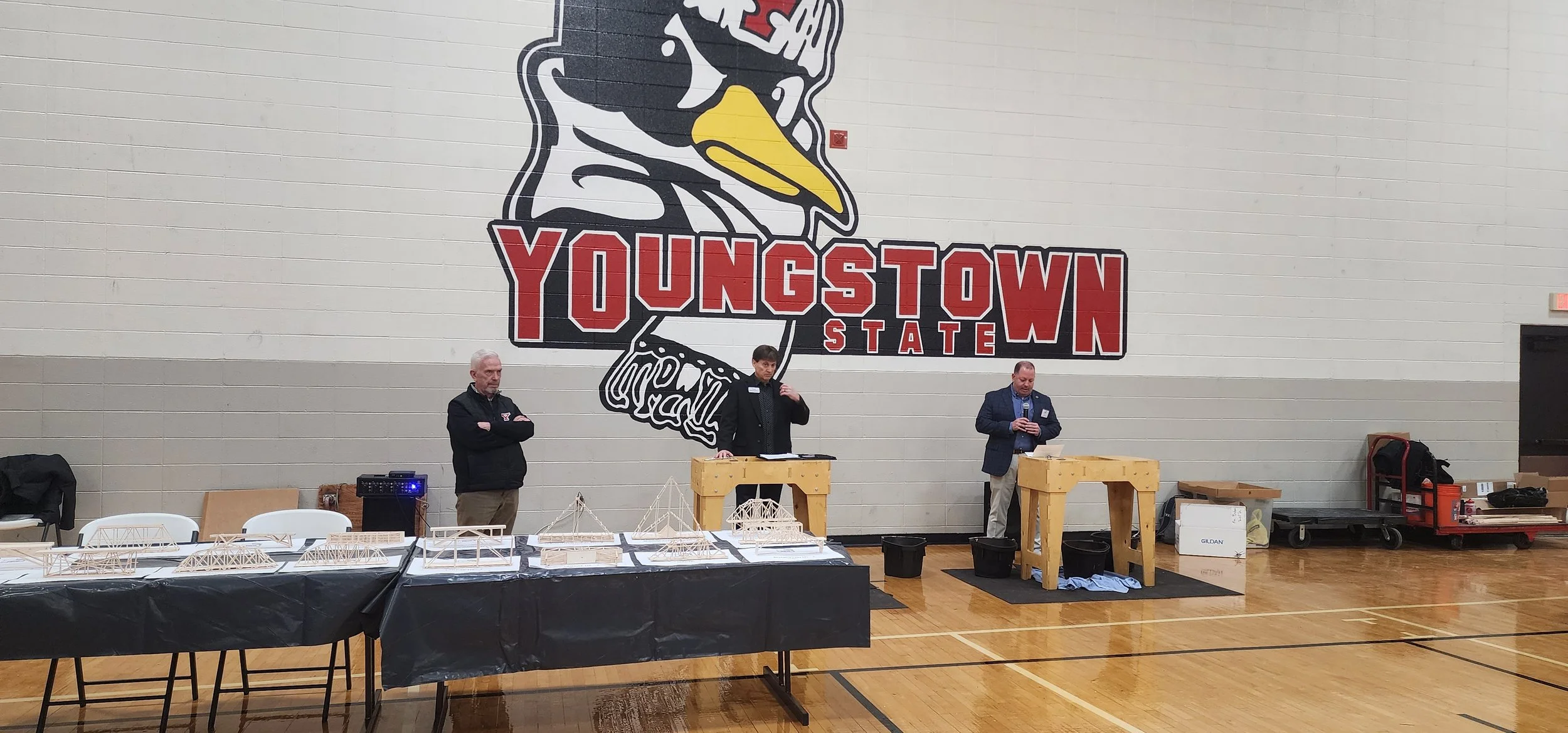 Three men are set up in front of a large wall with a graphic of a duck and the words "YUNGSTOWN STATE." They are standing behind tables with model bridges and building materials. One man on the right is speaking into a microphone, the middle man is a