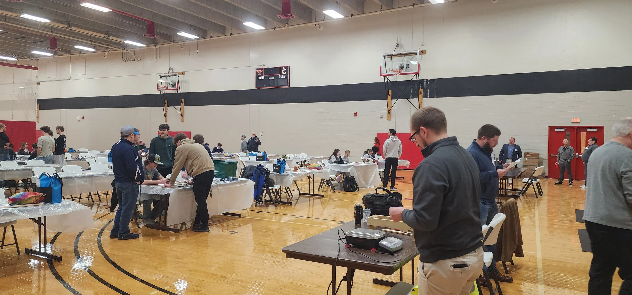 People gathered inside a gymnasium with tables set up, possibly for an event or a community activity. Some individuals are engaged in conversations or looking at papers, with a basketball hoop visible on the wall.