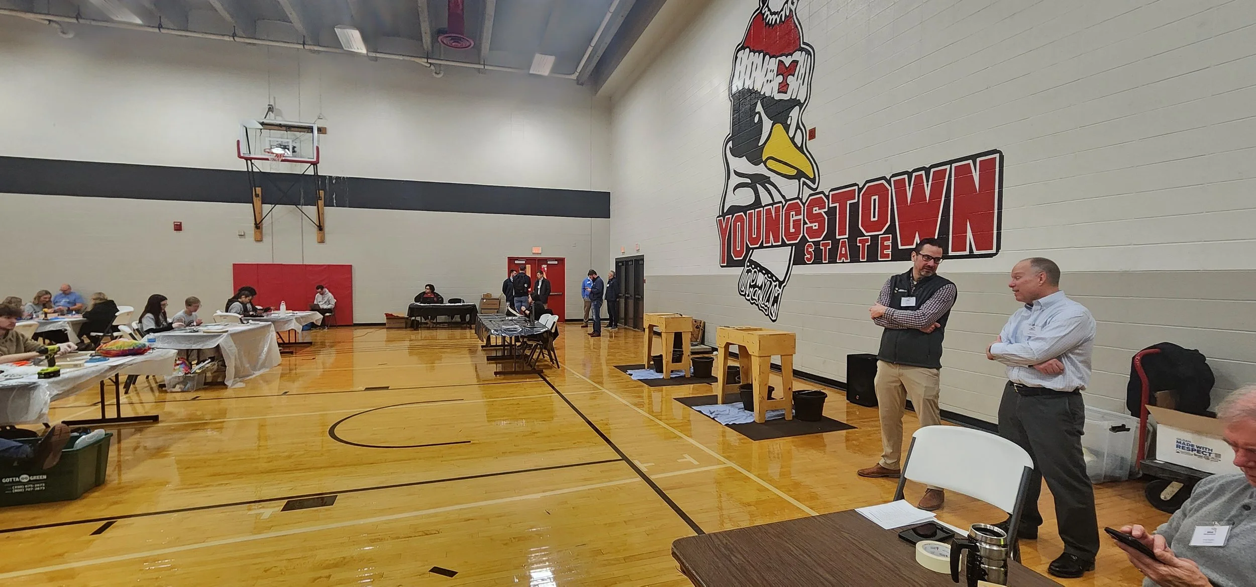 An indoor gymnasium with a large Youngstown State University logo painted on the wall. Several people are sitting at tables covered with white tablecloths, and some are standing and talking near the wall. There are booths and equipment set up for an 