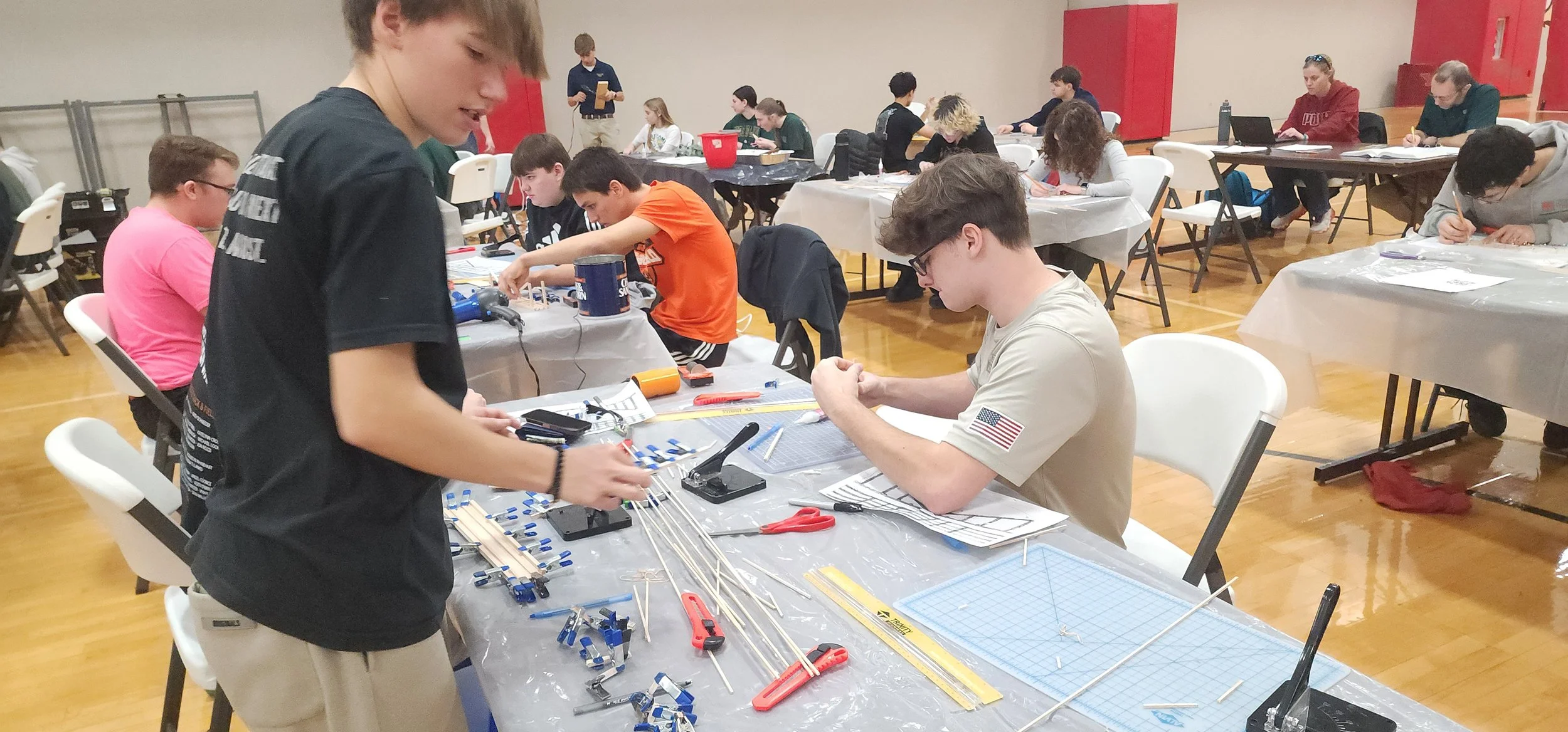 Students working on a engineering or technical project at tables filled with tools, wires, and model components in a classroom or workshop setting.