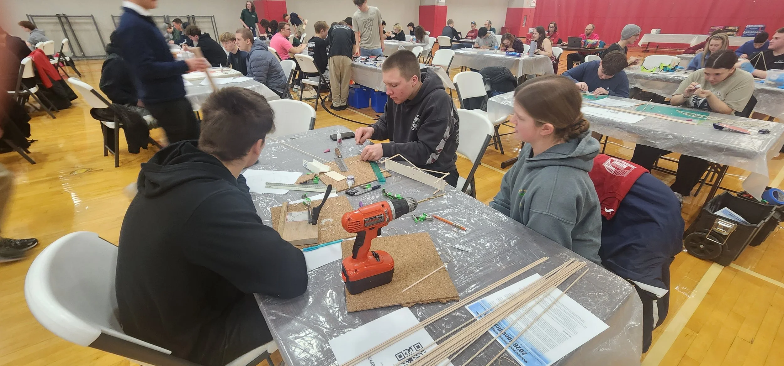 Students working on a project involving wooden sticks and tools at a classroom or workshop setting with many tables and people in the background.