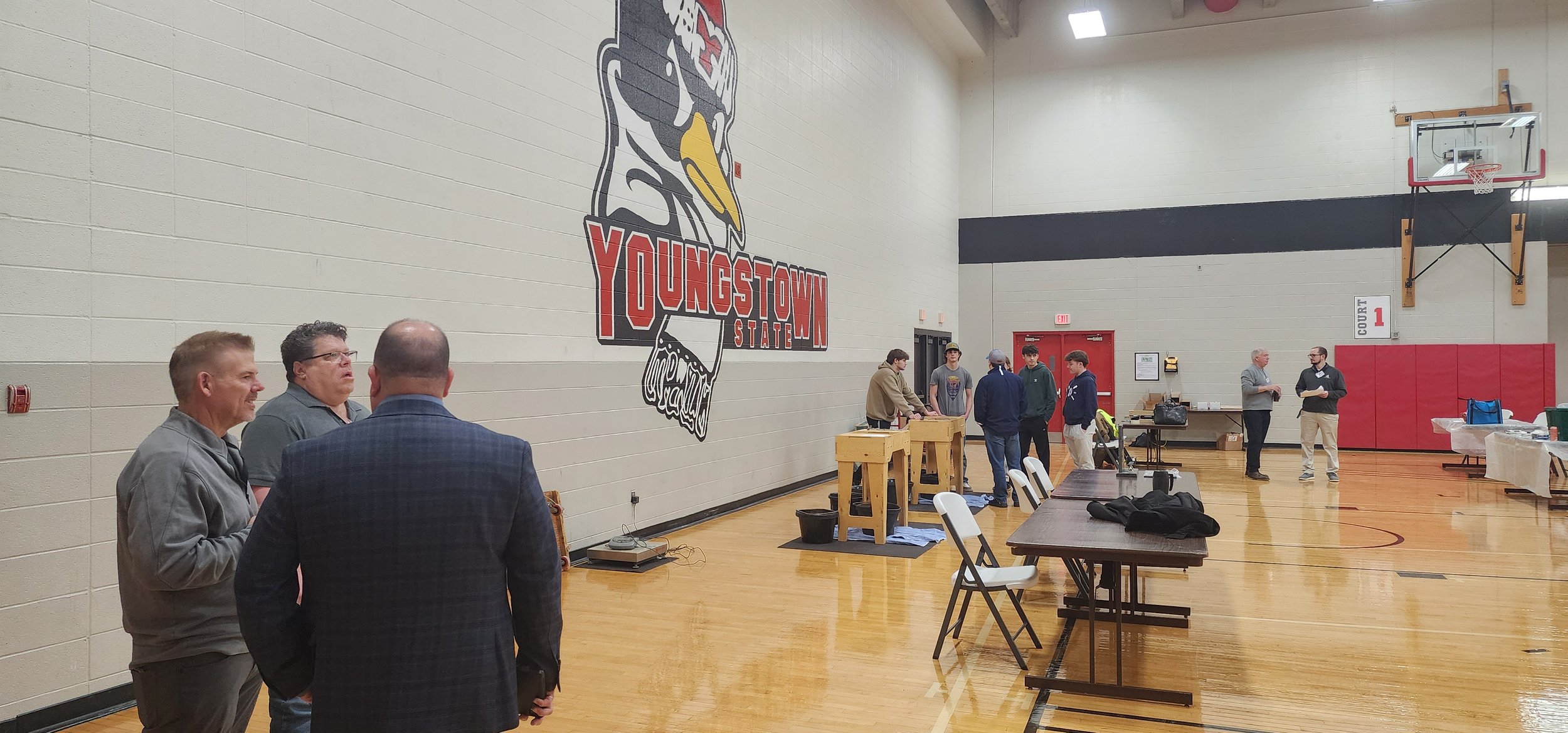 A gymnasium with a large mural of a youngstown state logo on the wall. Several people are gathered, some talking and others working at tables or standing. There are tables and chairs set up for an event.
