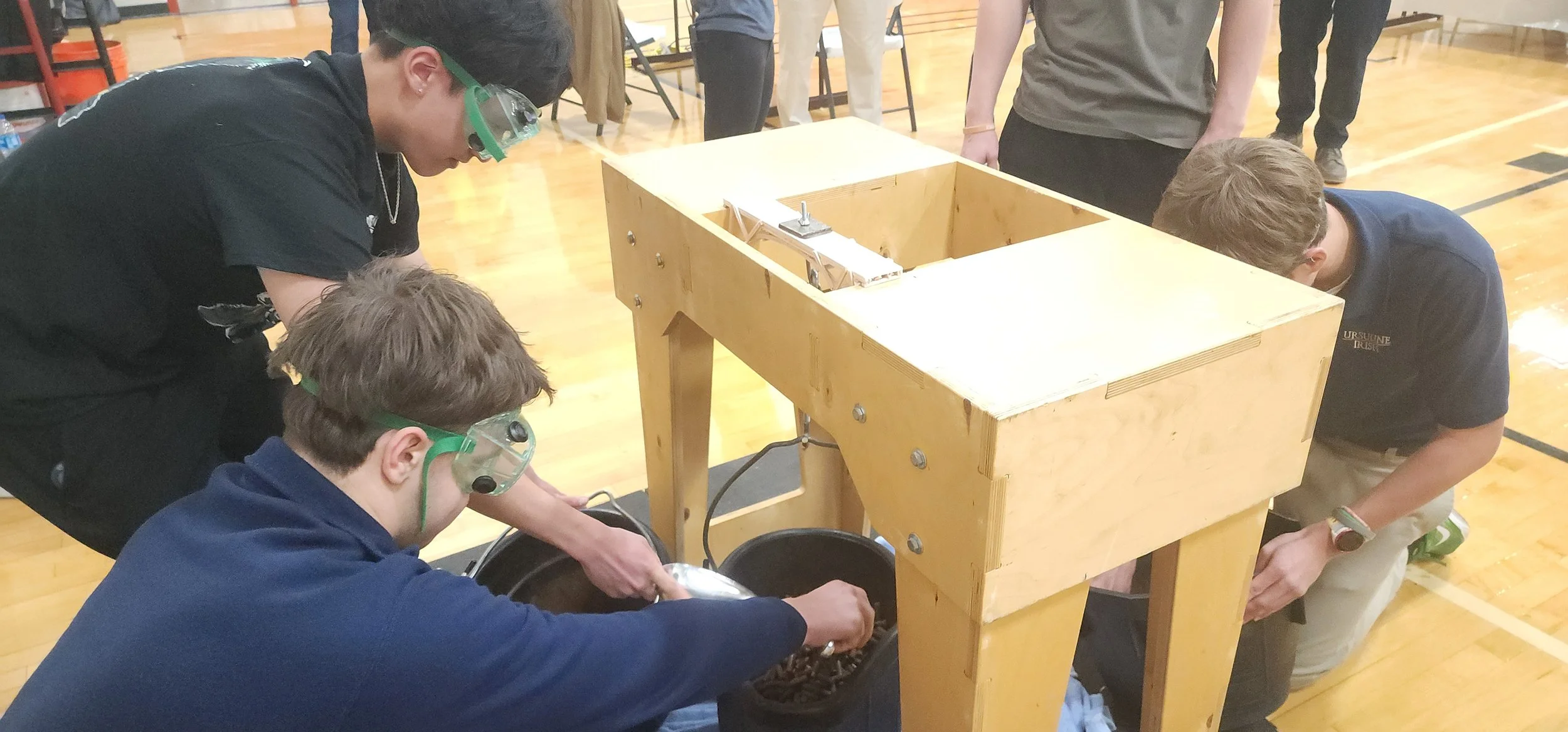 Students in goggles working with a mechanical device on a wooden table in a gymnasium.