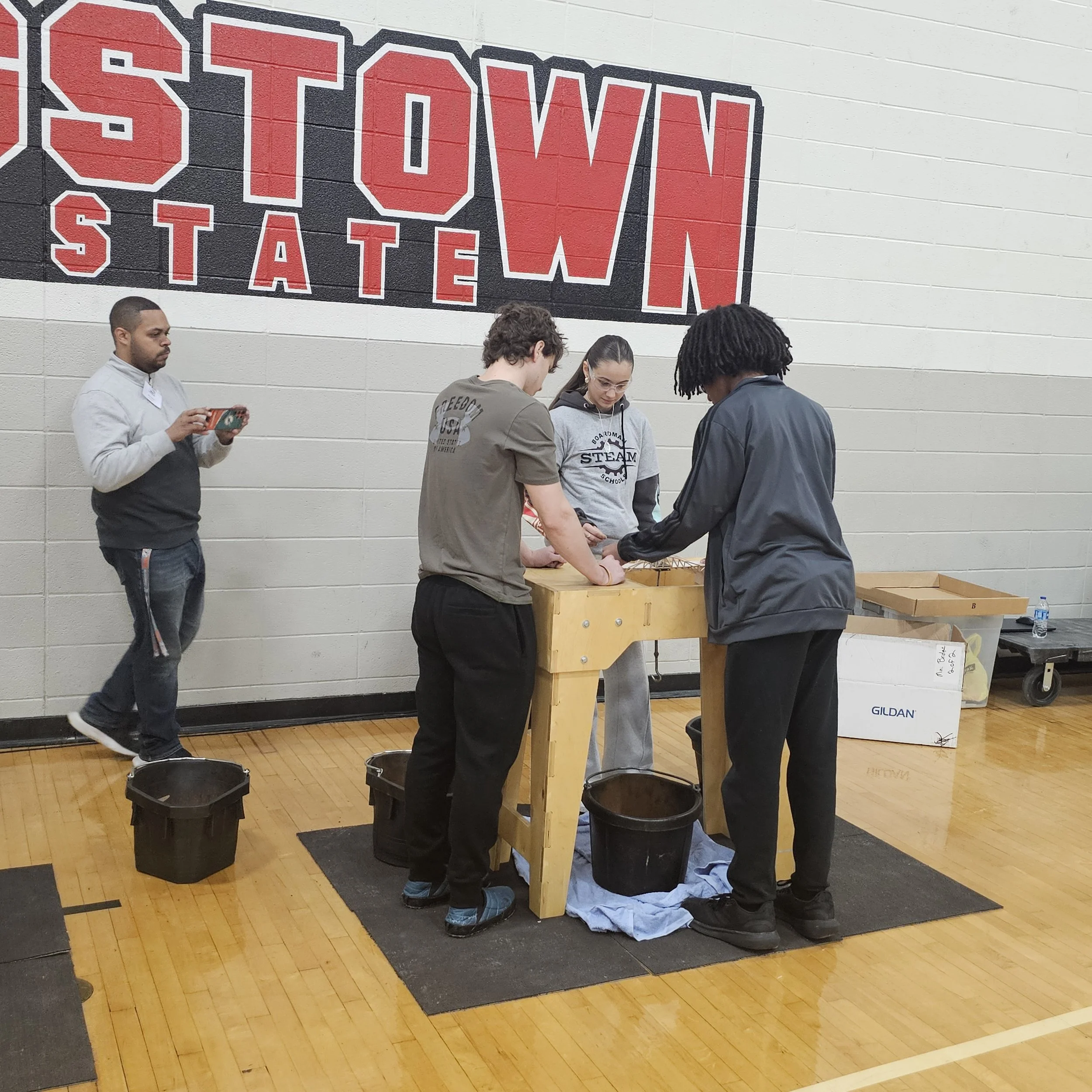 People participating in a tabletop activity at an indoor event, with a large red and black 'STATE' sign on the wall behind them.
