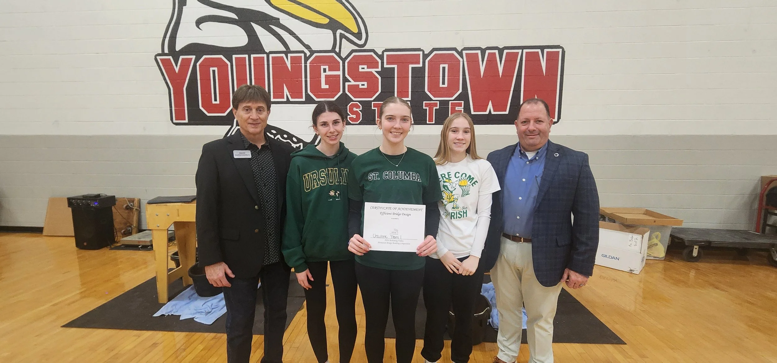 Group of five people standing in front of a wall with a 'Youngstown' logo, three young women in the center holding a certificate of achievement, flanked by two adults, in a gymnasium.