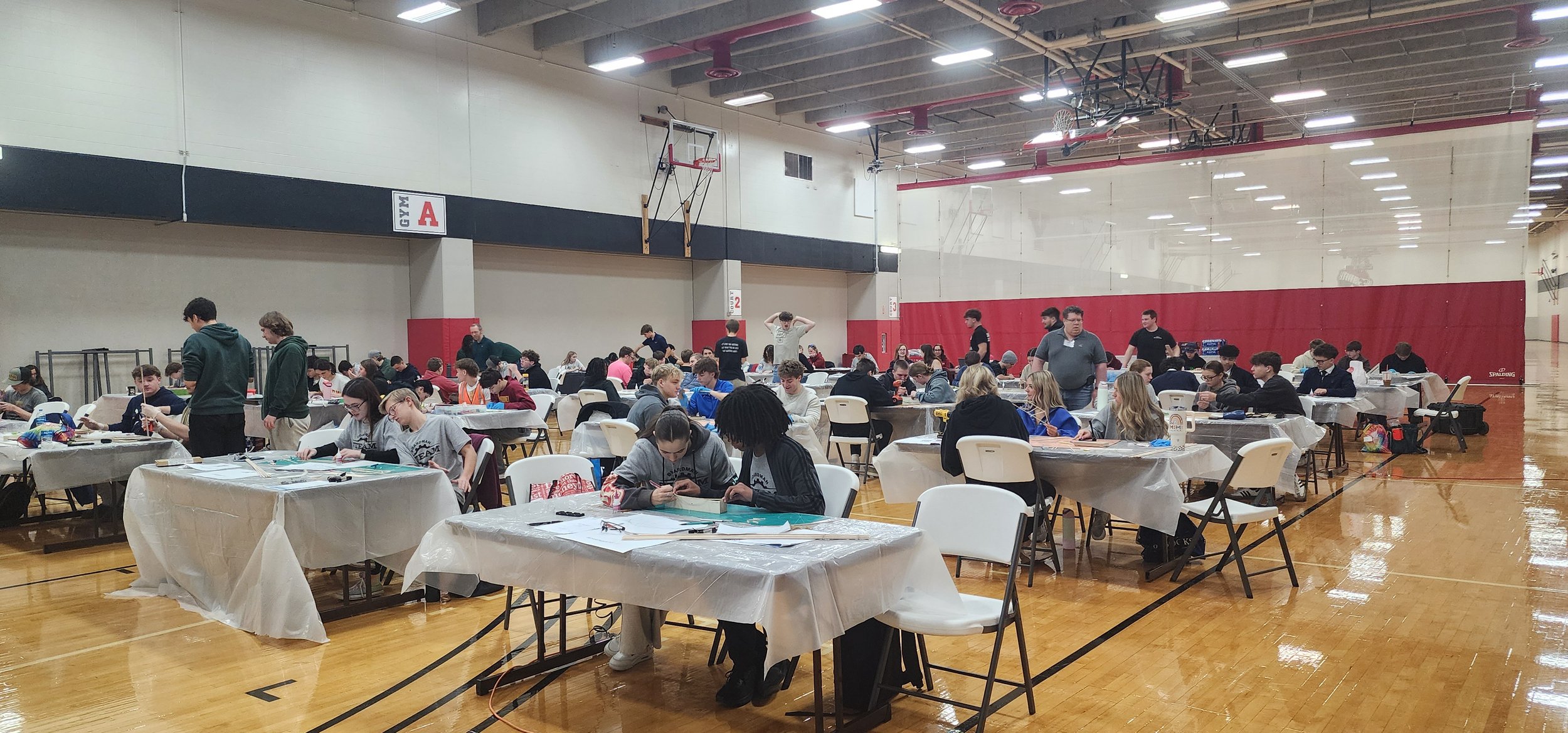 A large indoor gymnasium filled with many people, mostly young students, seated at tables covered with white tablecloths, engaged in arts and crafts activities. Some individuals are standing and walking around. The gym has a high ceiling with bright 
