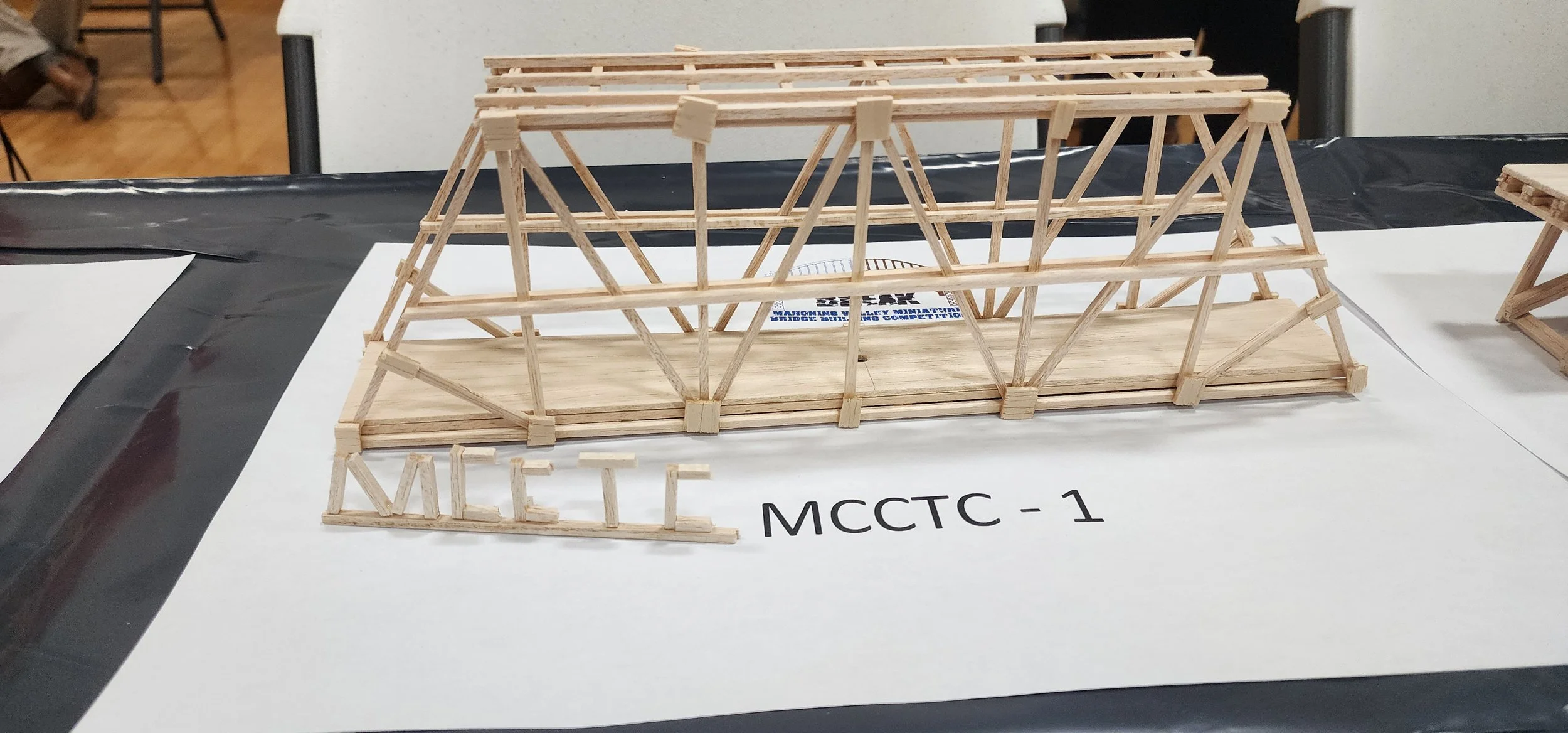 A miniature wooden structure resembling a building frame, placed on a white display board with the label MCCTC-1. The structure has multiple interconnected wooden pieces forming a truss-like framework.