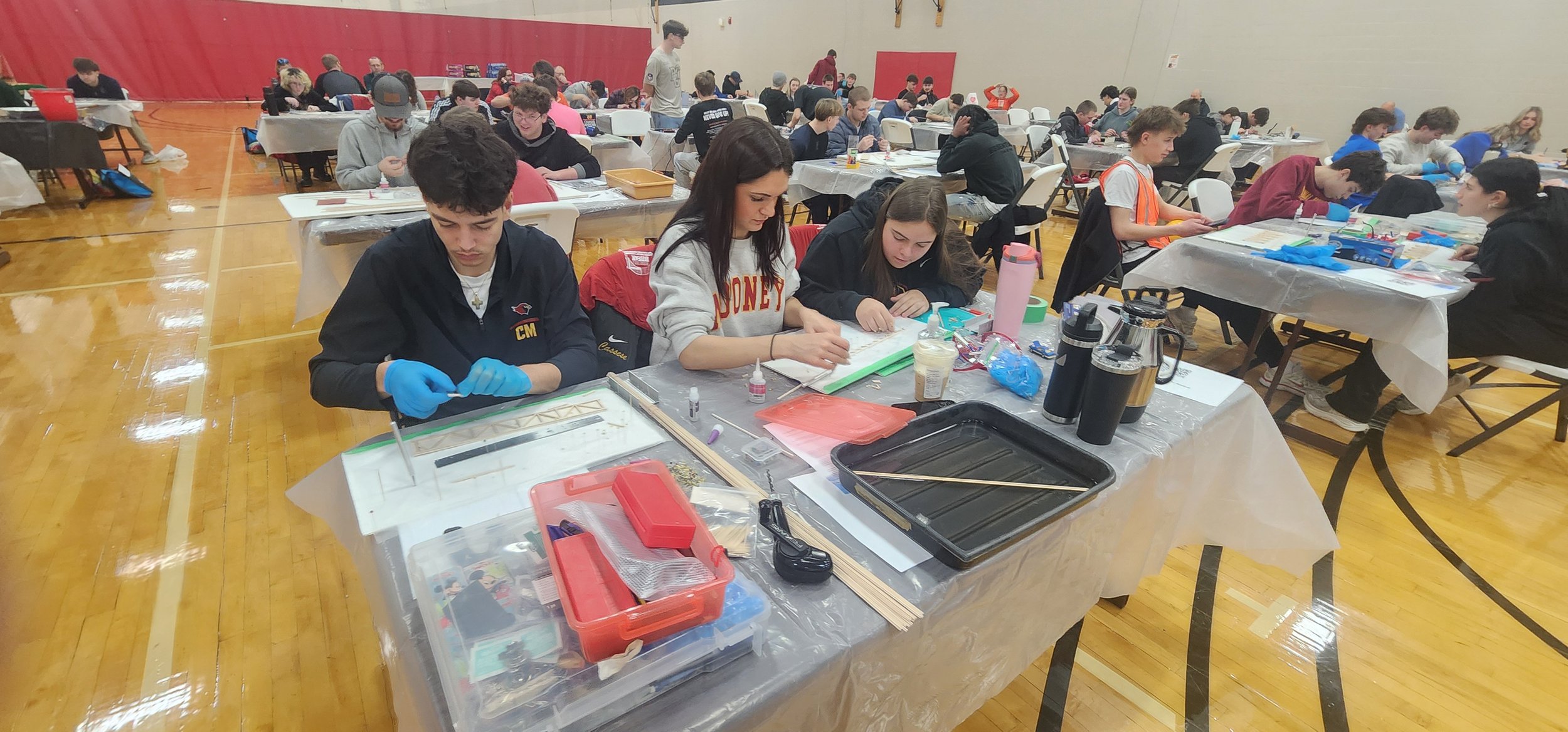 Students seated at tables in a gymnasium, working on science projects with various materials, some students wearing gloves and safety gear, and others engaged in discussions.
