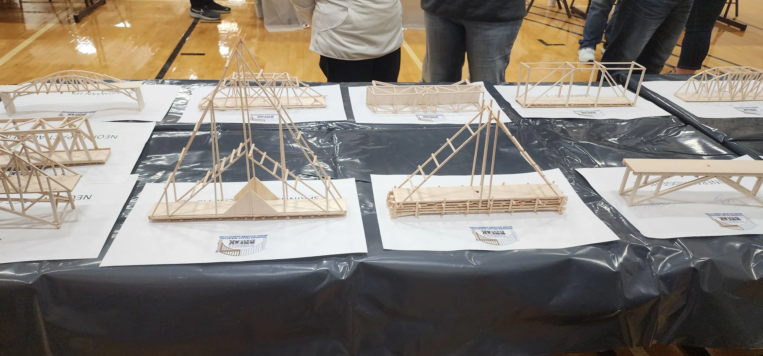 Display of small wooden architectural bridge models on black tables, with people viewing them in an indoor gymnasium.