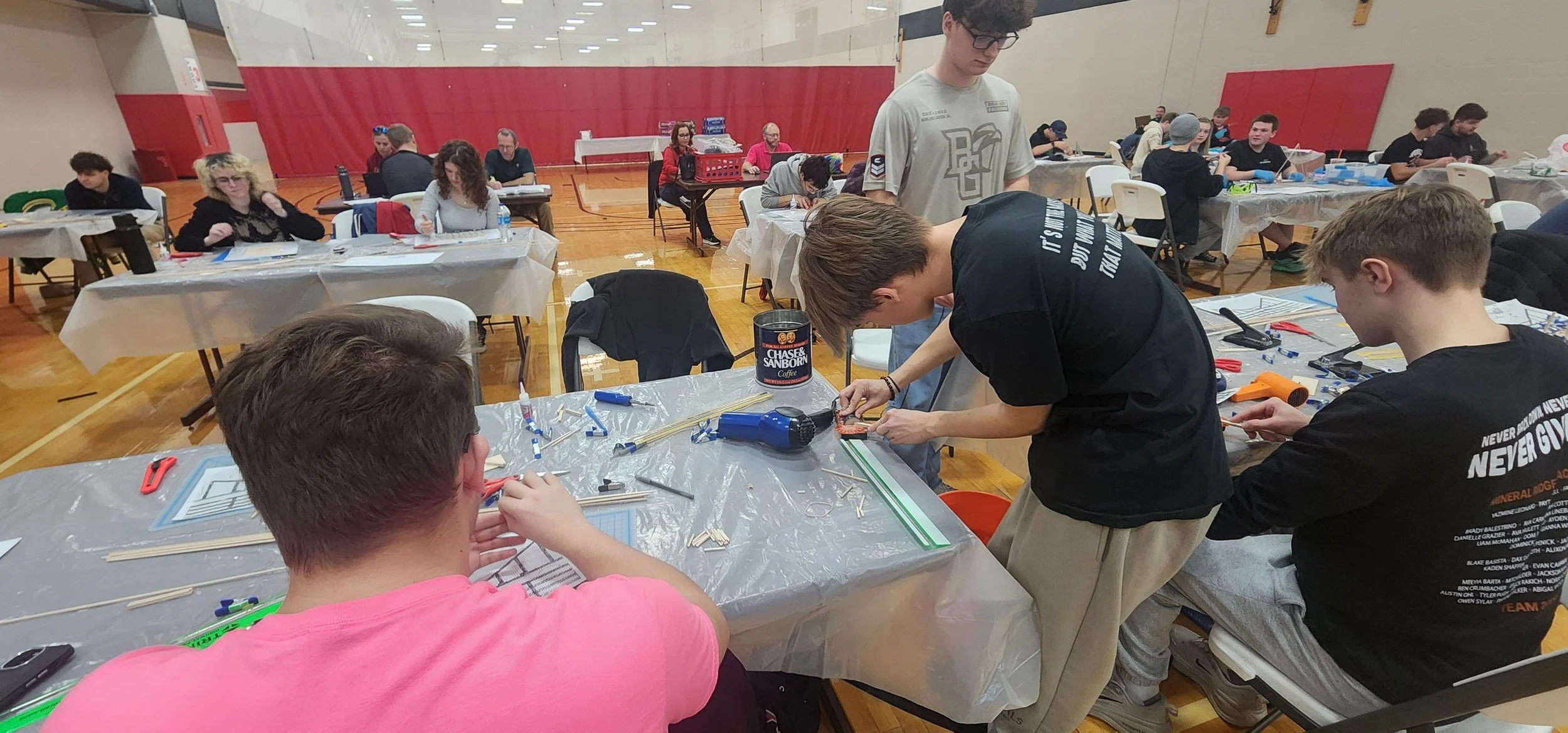 Group of people working on a robotics project at tables covered with plastic. Some are assembling parts, others are observing or taking notes, in a gymnasium.