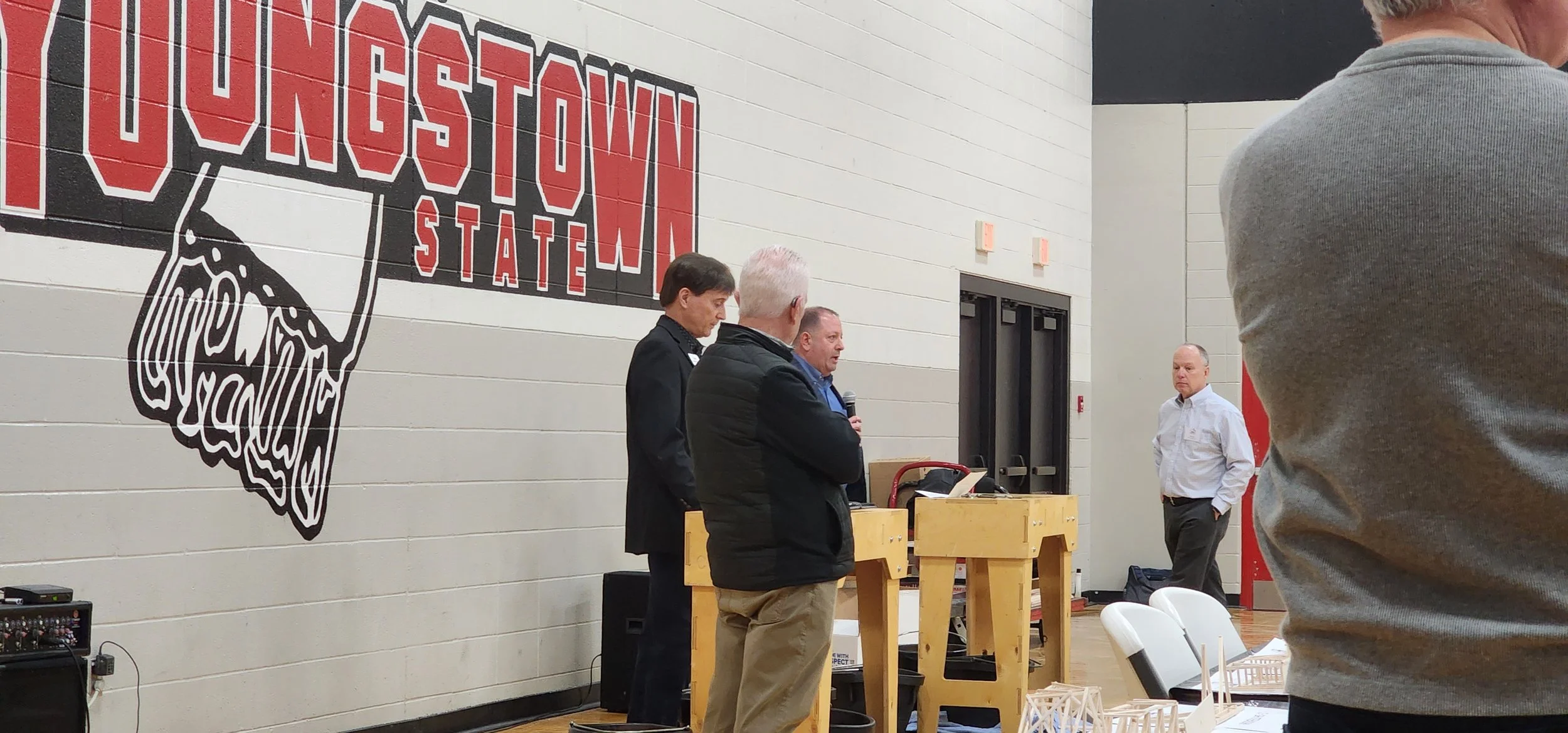People standing and speaking at an indoor event with a large wall sign reading 'Huntington School' and a depiction of the state of West Virginia.