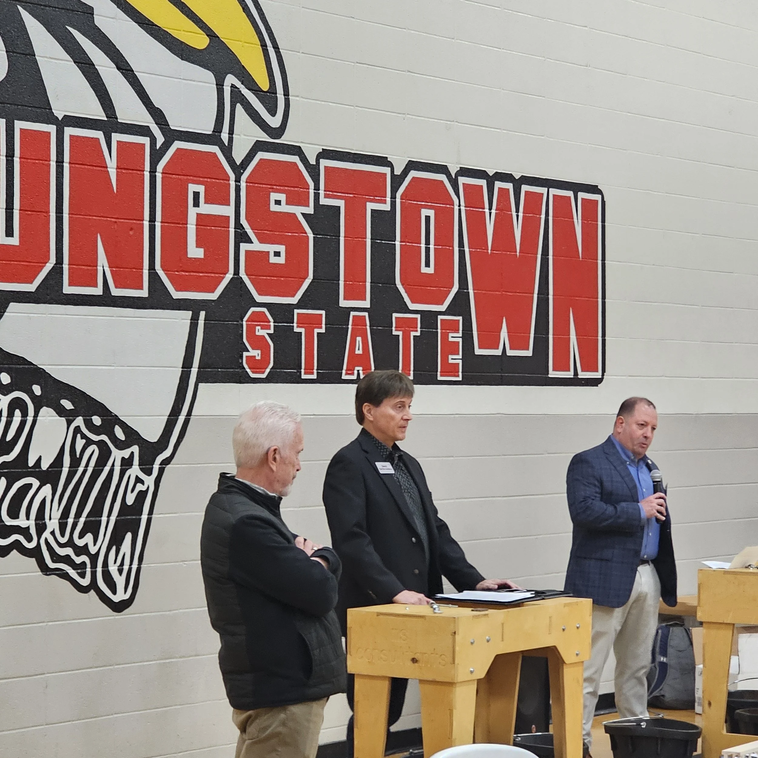 A group of people standing behind a wooden podium during a presentation or ceremony in a gymnasium or similar venue. The background features a large lion logo painted on the wall, with the word 'STATES' visible. There are three men in the image, one 