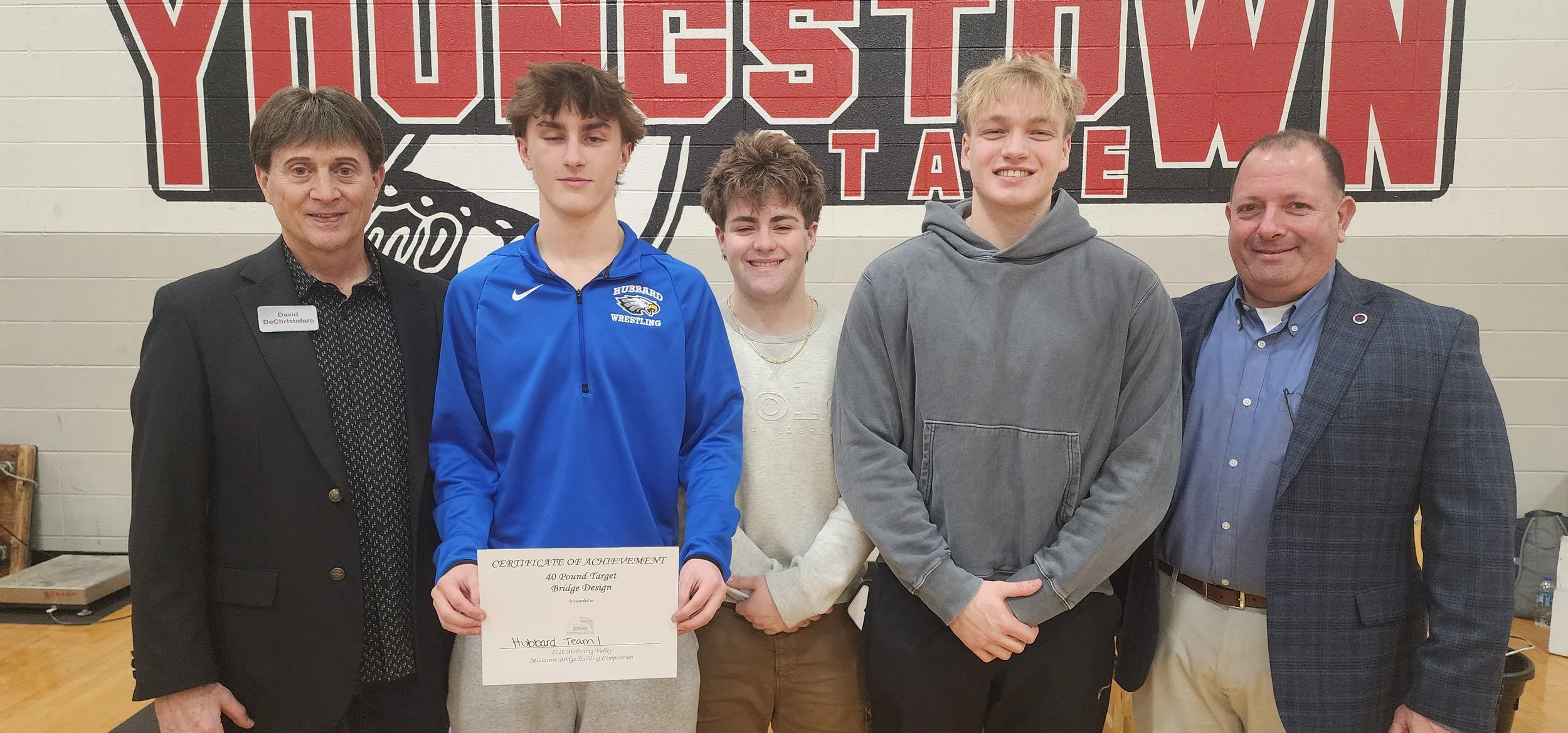 Group of five people standing indoors in front of a wall with school logo. One person in the center holds a certificate. Two are adults, three are young males. All are smiling or with neutral expressions.