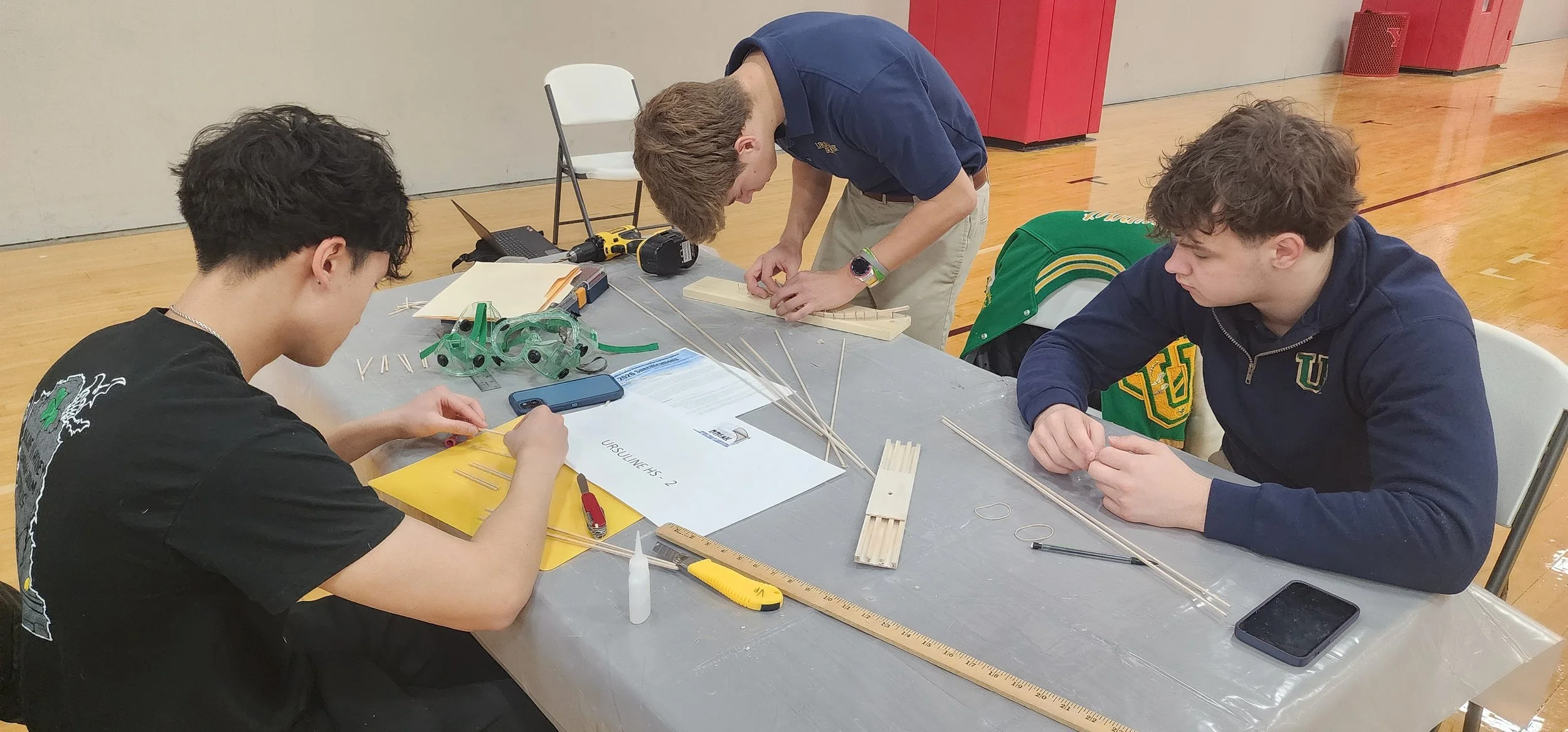 Three young men working on a project at a table in a gymnasium. They are surrounded by various tools and materials, including wooden sticks, rulers, and a tape measure.