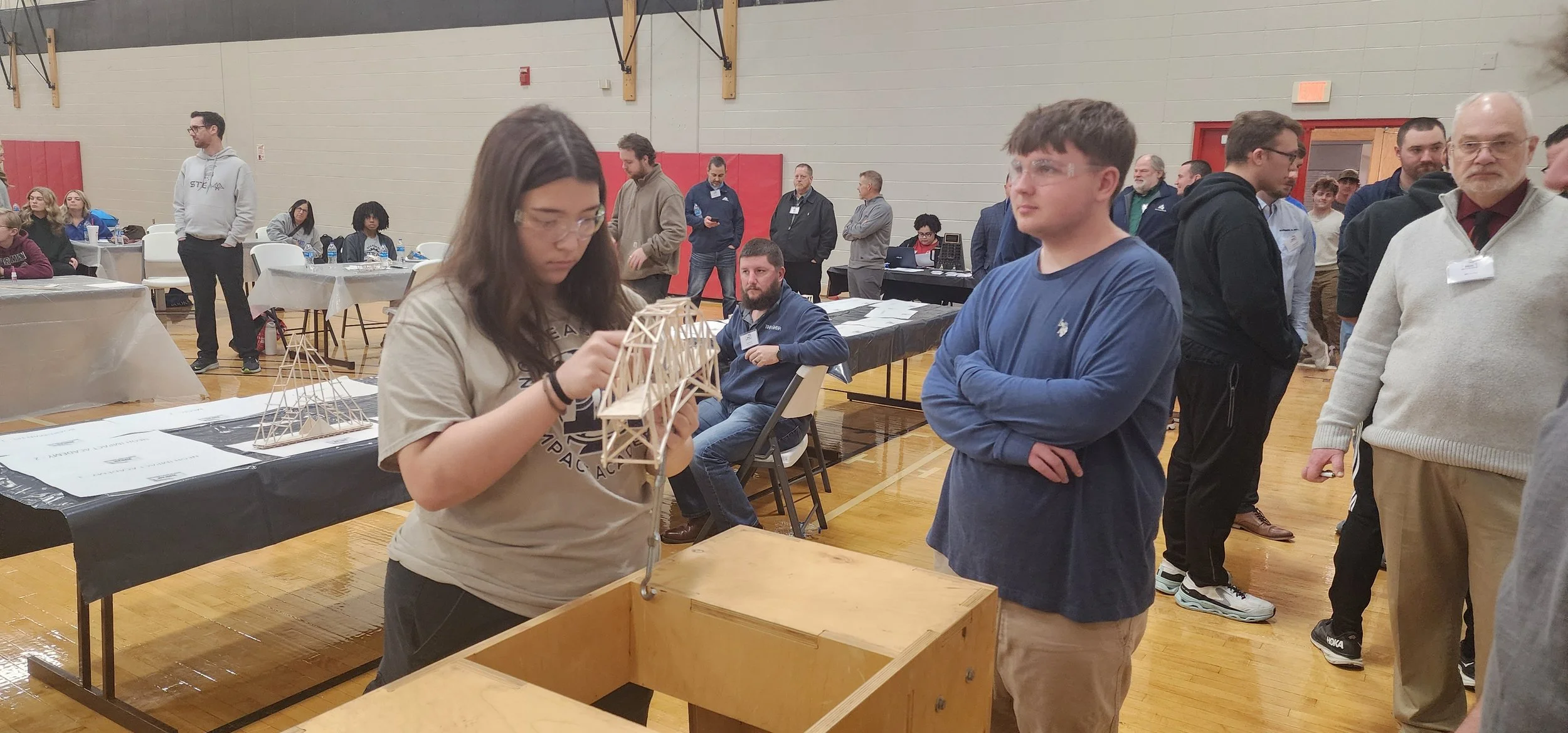Young woman working on a wooden bridge model at a table, with a young man observing her, in a busy indoor event hall.