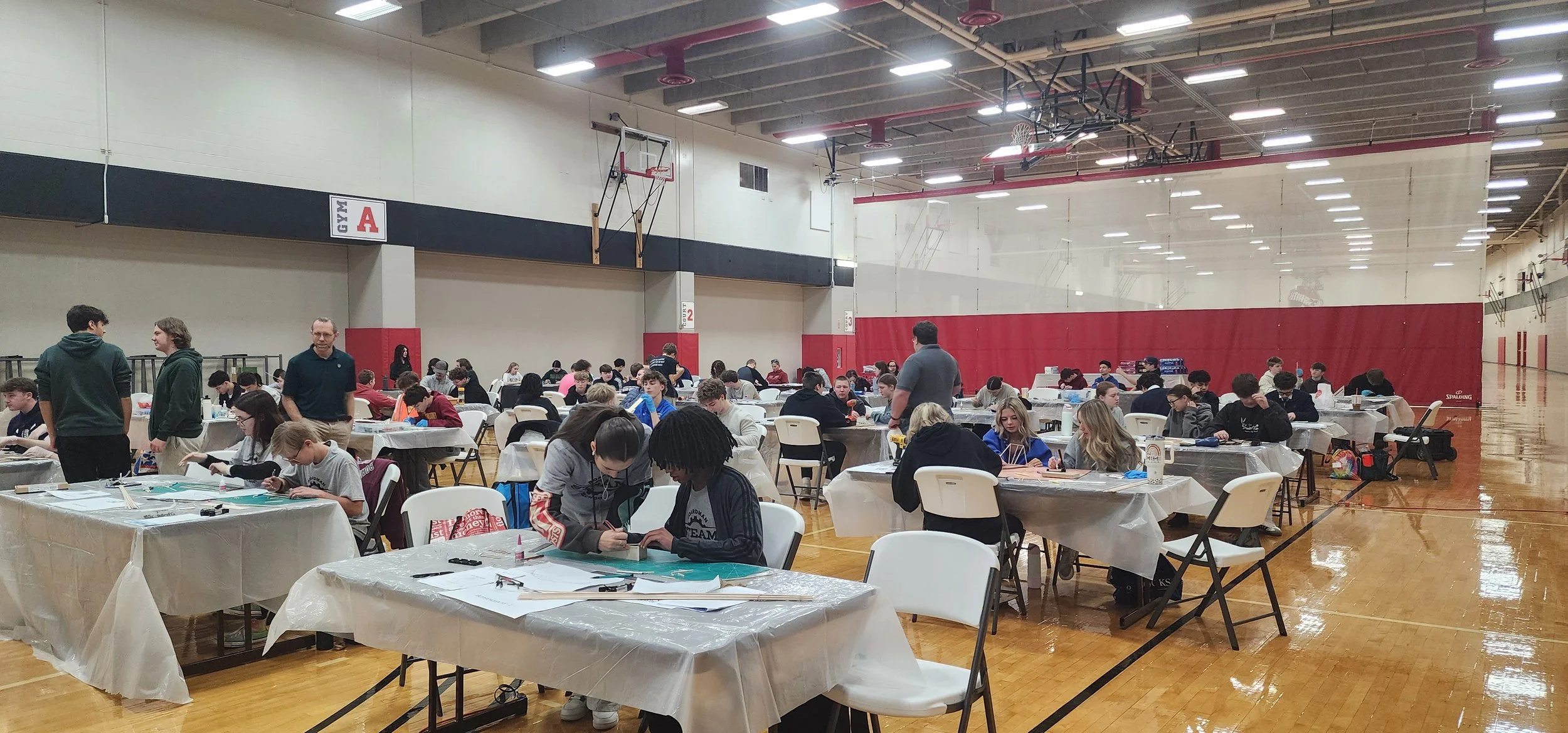 A large gymnasium filled with students seated at tables covered with plastic tablecloths, engaged in various activities, with a few adults walking around. The gym has high ceilings, basketball hoops, and wood flooring with red and cream walls.