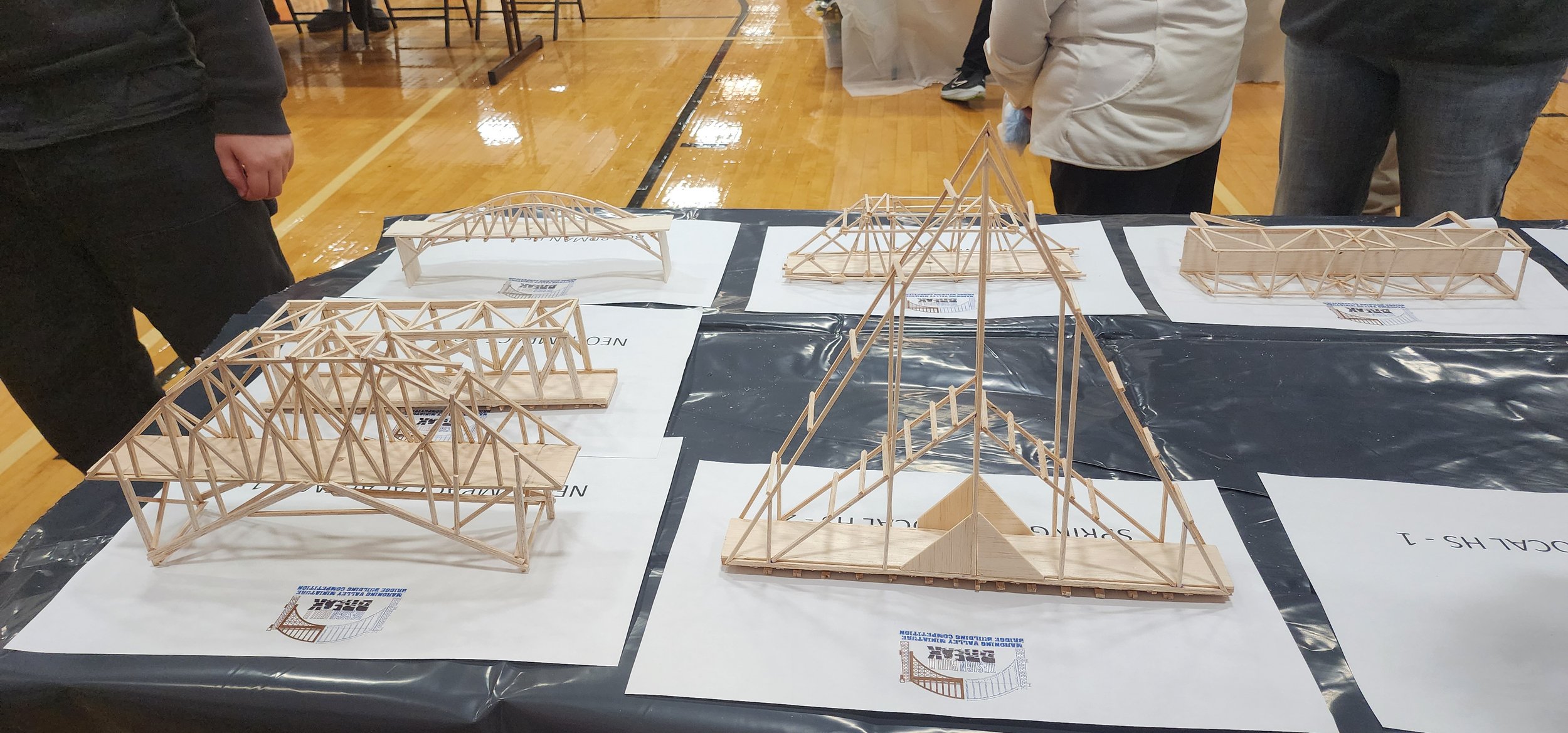 Various small wooden models of bridges and structures displayed on a table at a model building competition in a gymnasium.