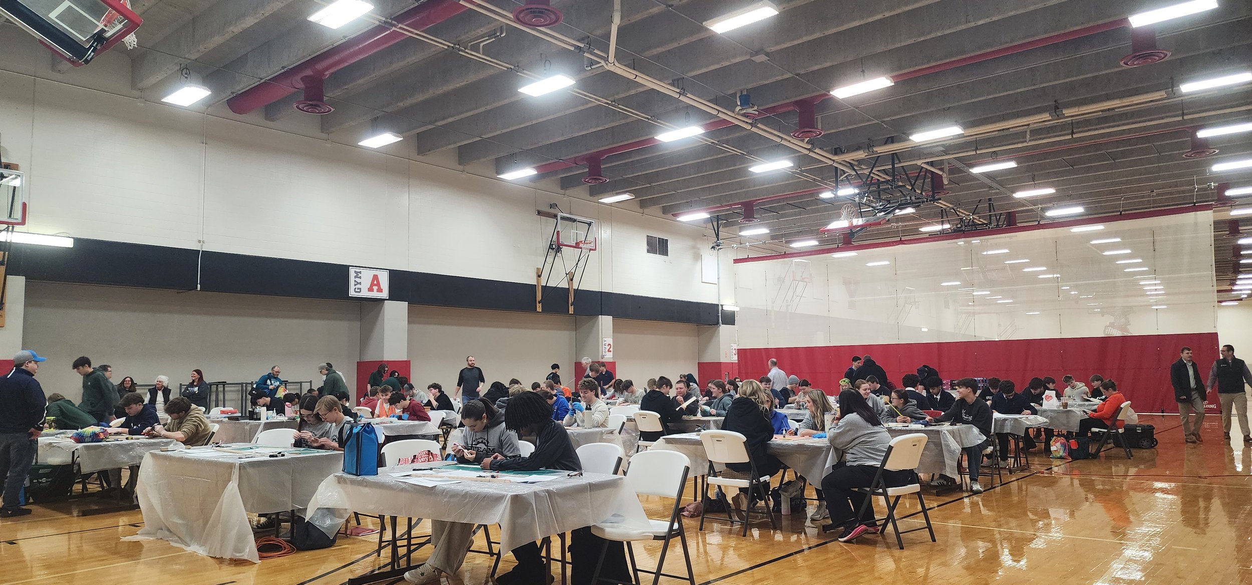 People sitting at tables in a gymnasium, some standing and walking, with basketball hoops and a large mirror on the wall.