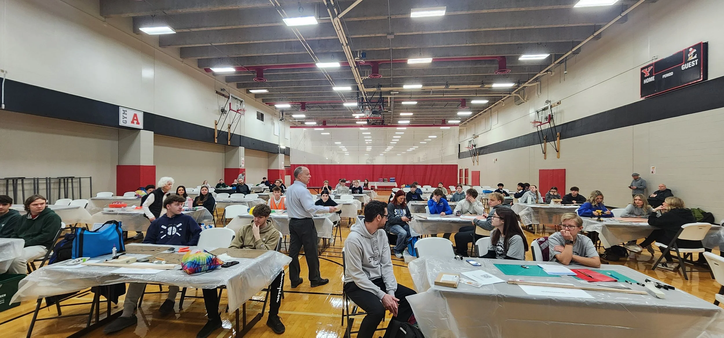 A large gymnasium with students seated at tables covered with papers and materials, a man standing and talking to the students, basketball hoops on the walls, and an electronic scoreboard overhead.